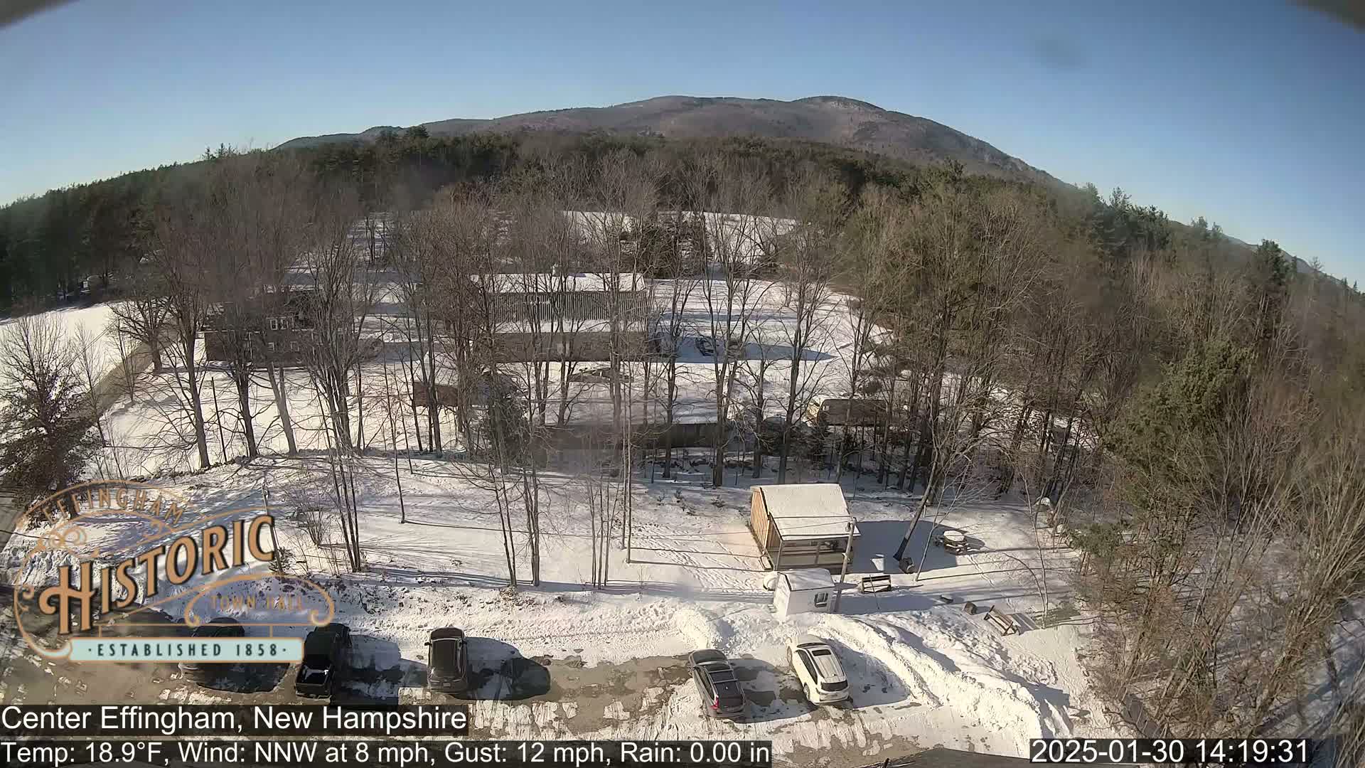 An aerial view shows a snow-covered parking lot with several vehicles, a small building, and a larger building surrounded by bare, leafless trees under a clear, sunny sky.