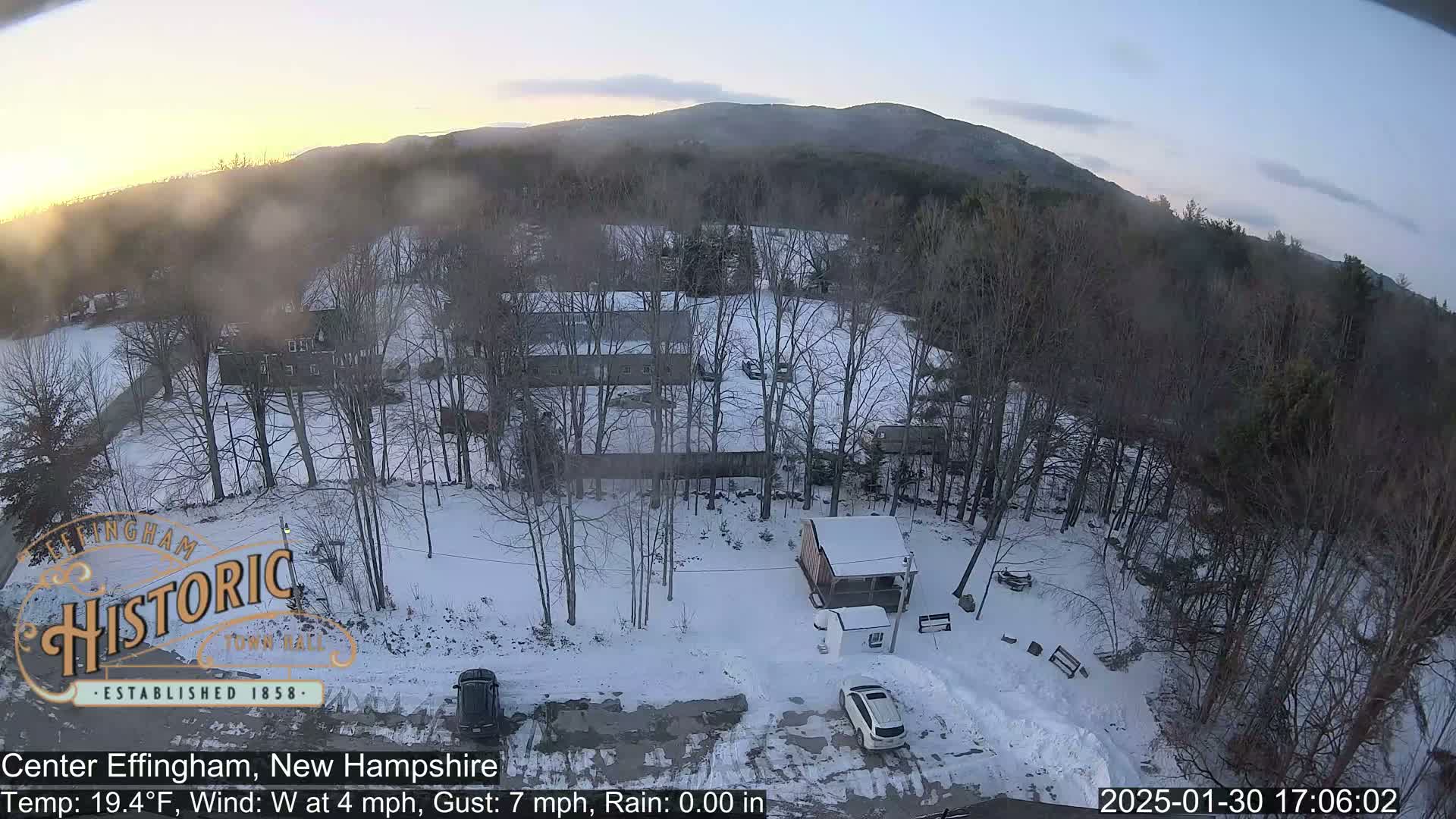 A snow-covered landscape with bare trees, a few buildings, and two vehicles in a parking area, under a twilight sky.