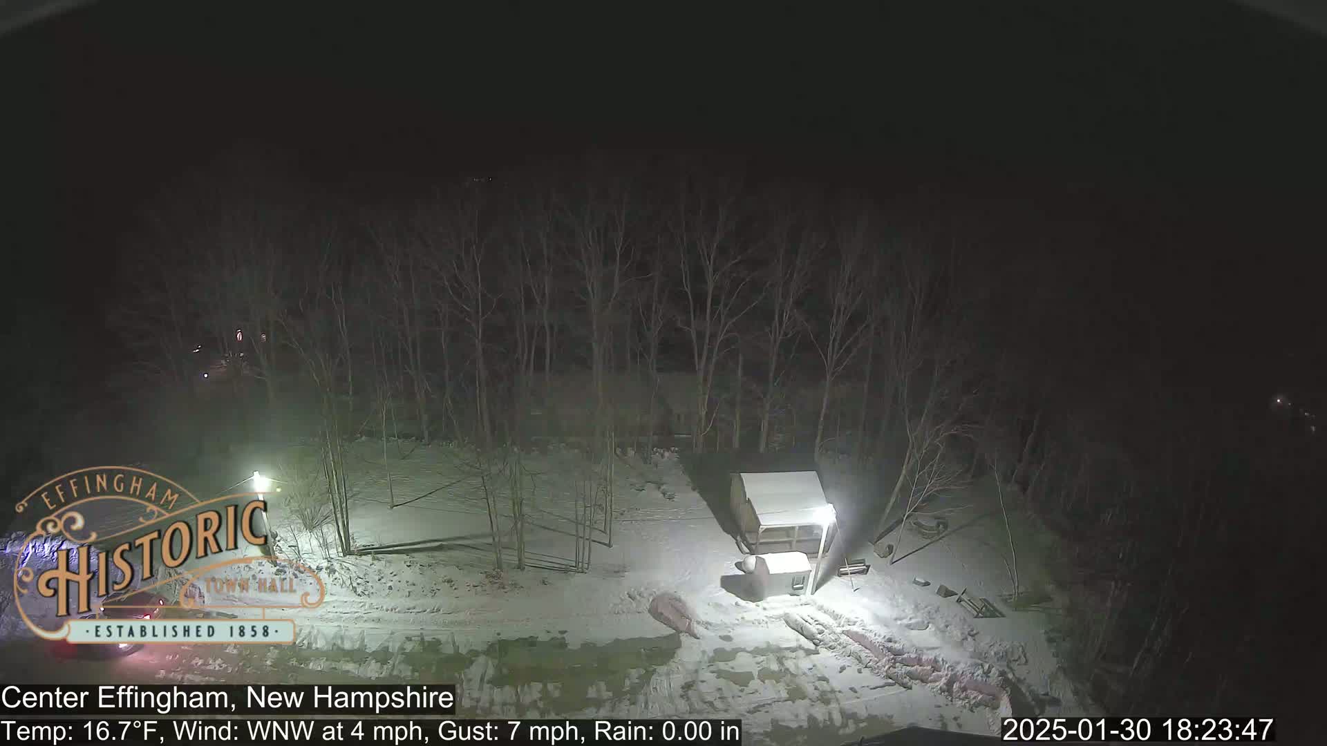A nighttime, snow-covered scene shows a small shed and bare trees under a dark sky.
