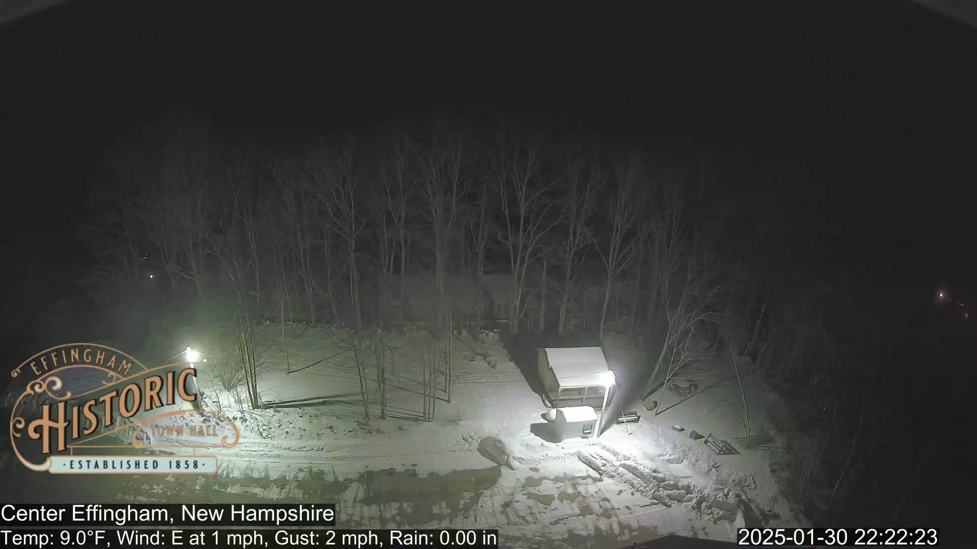A snow-covered area at night, lit by artificial lights, shows a small structure and bare trees under a dark sky.