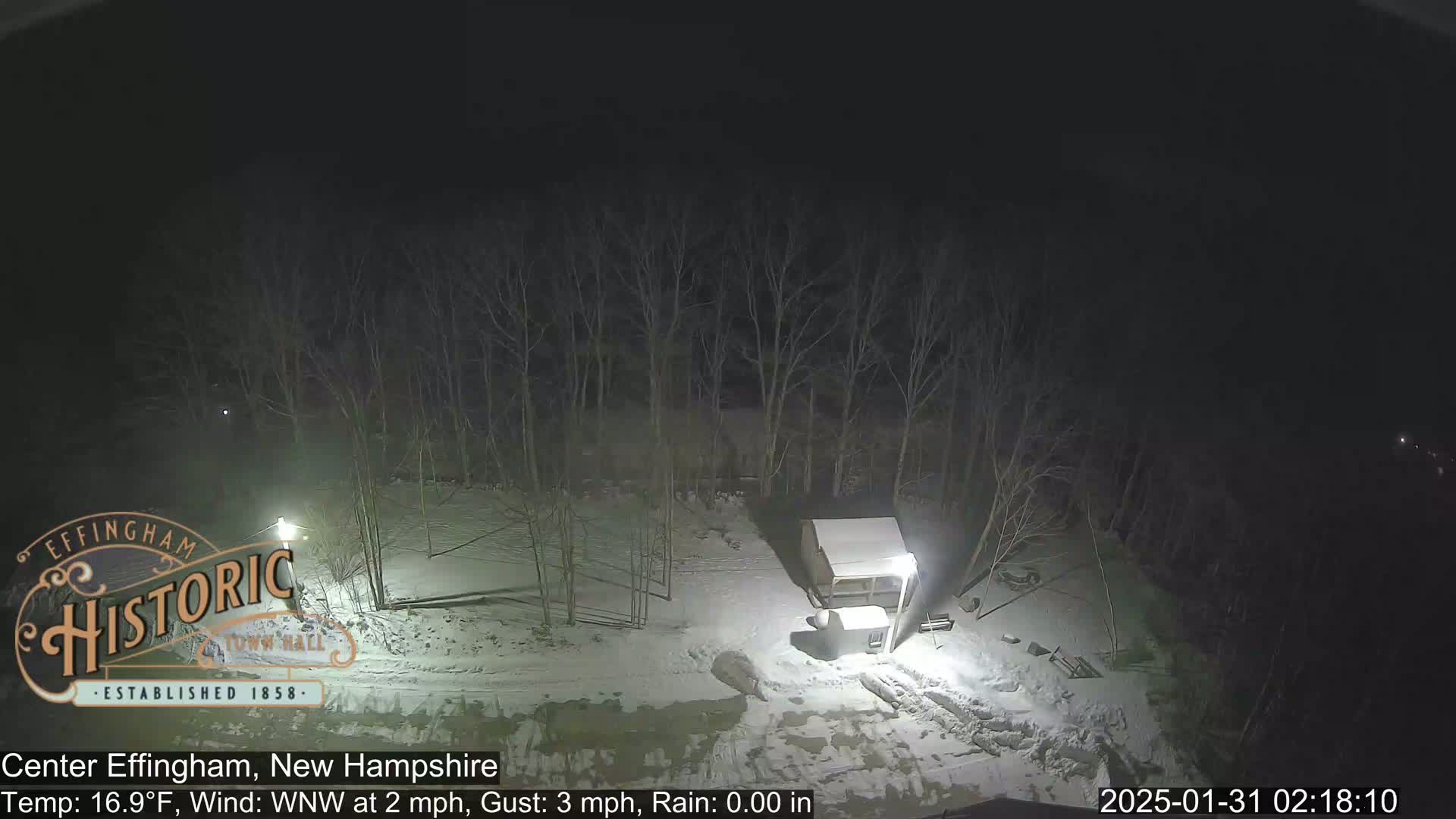 A nighttime, snow-covered scene shows a small structure and bare trees under a dark sky.