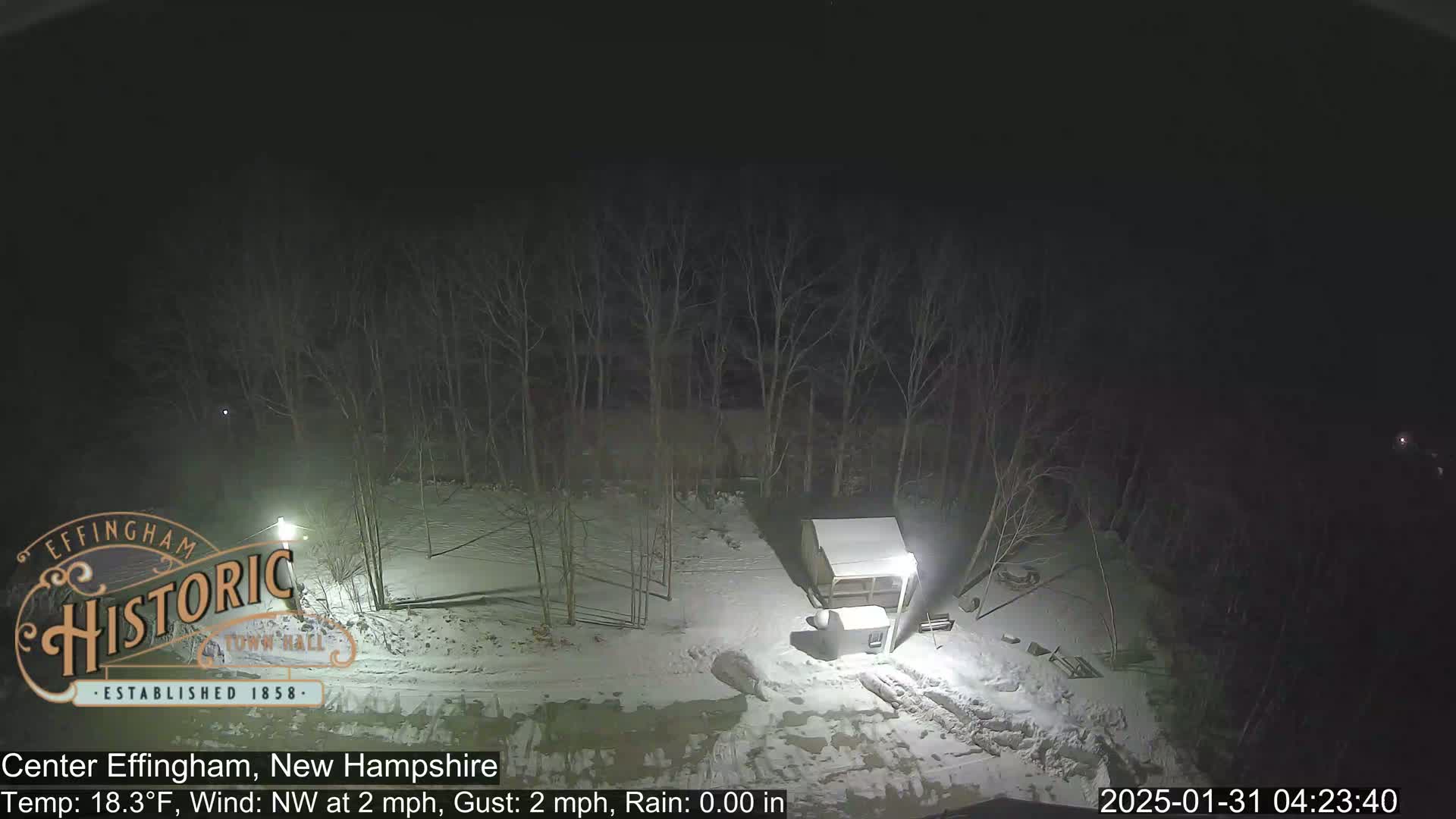 A snow-covered, nighttime scene shows a small shed and bare trees under a dark sky.