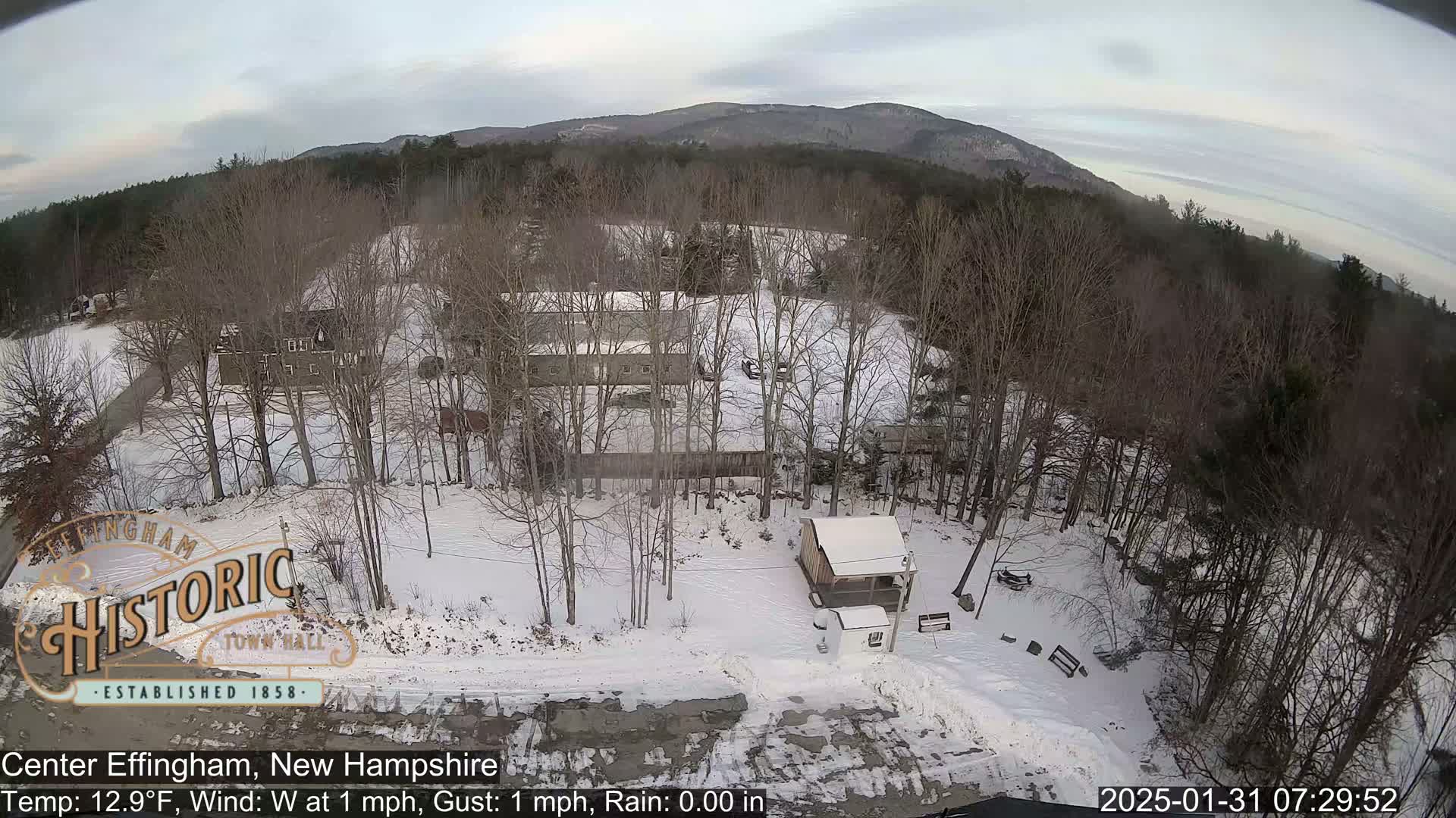 A snow-covered landscape with bare trees, several buildings, and a small structure, viewed from an aerial perspective under a cloudy sky.