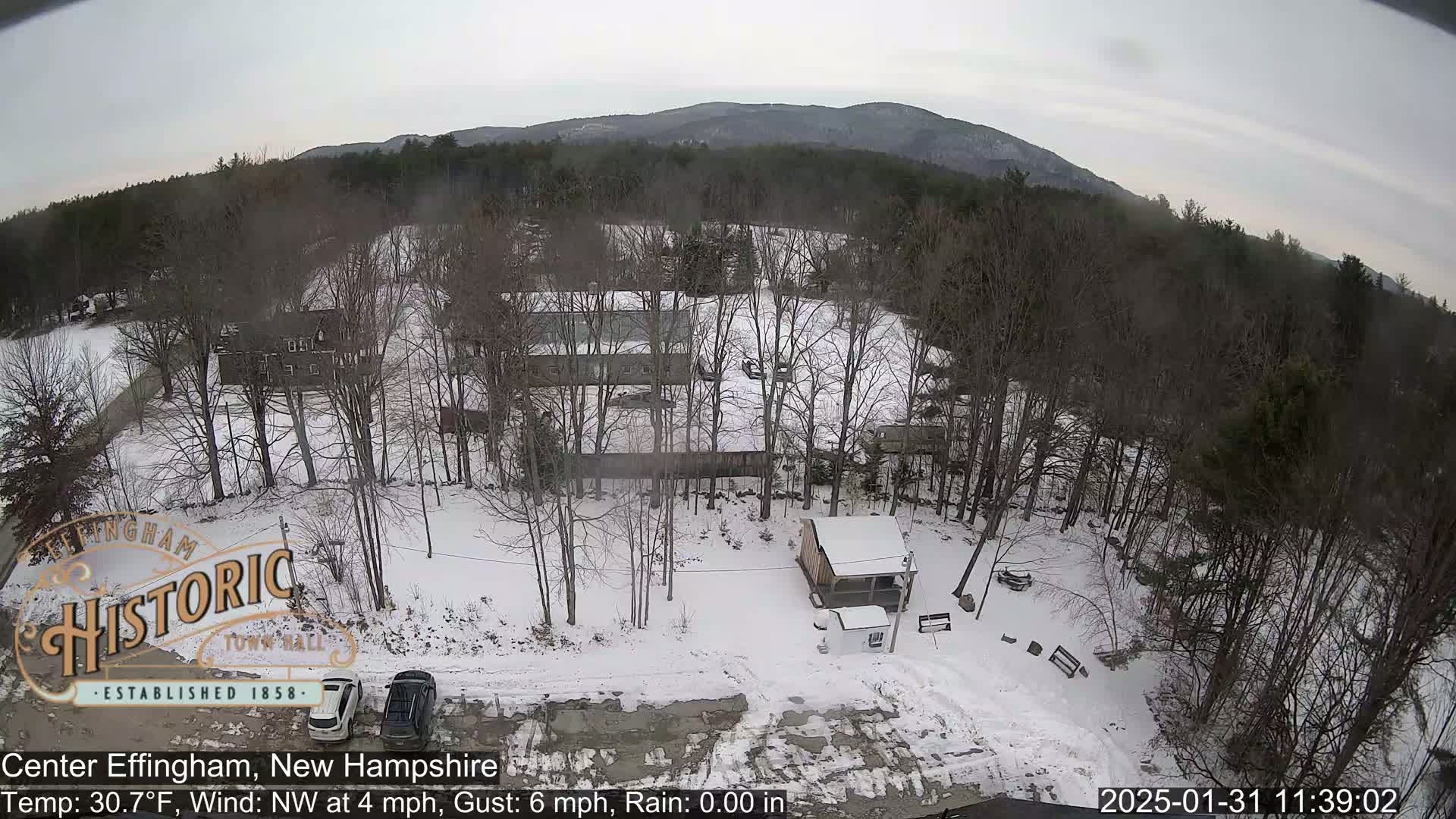 A snow-covered landscape with bare trees, several buildings, and two parked cars, viewed from above under an overcast sky.