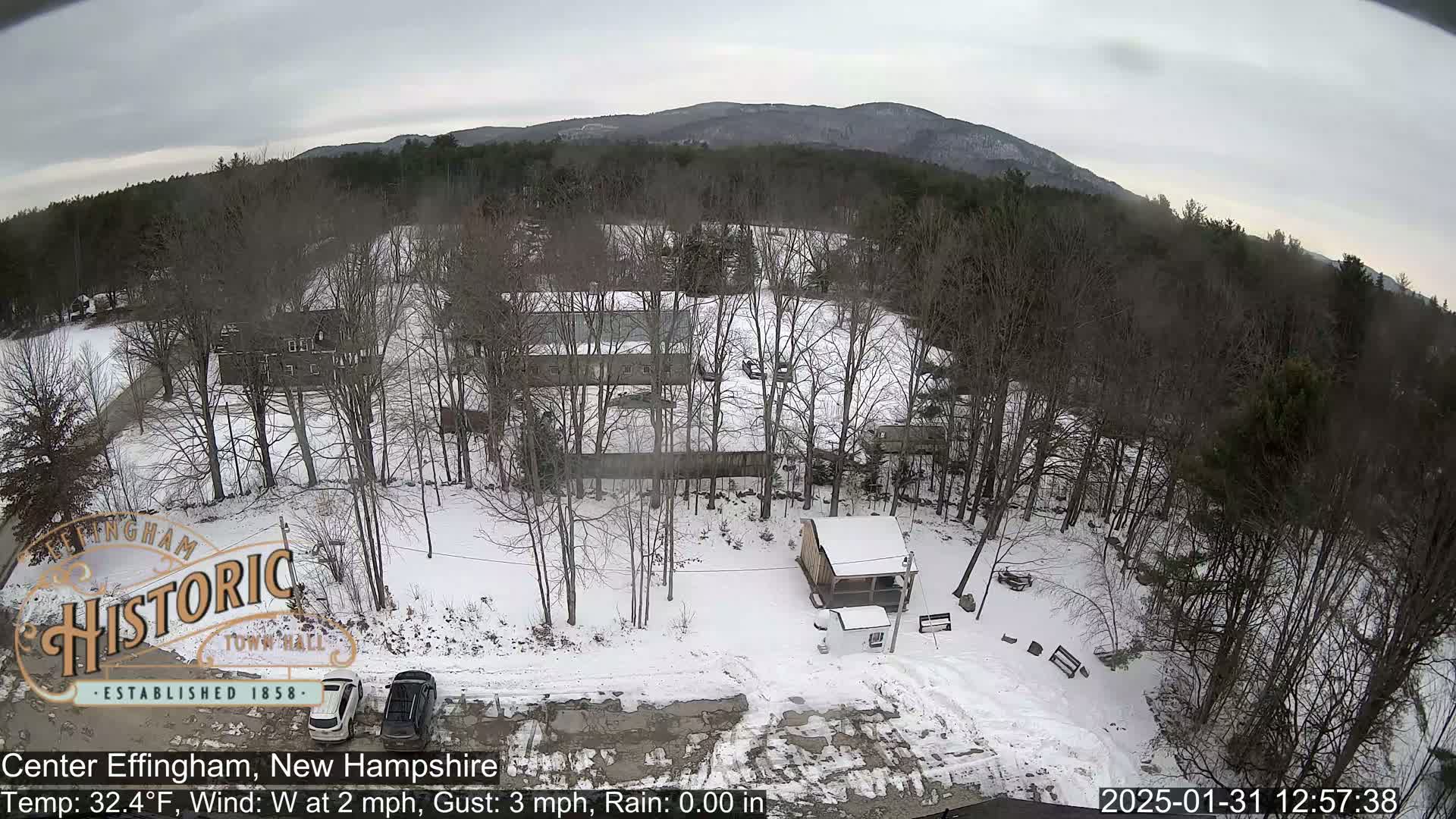 A snow-covered landscape with bare trees, a small building, and two cars in a parking lot, viewed from above under an overcast sky.