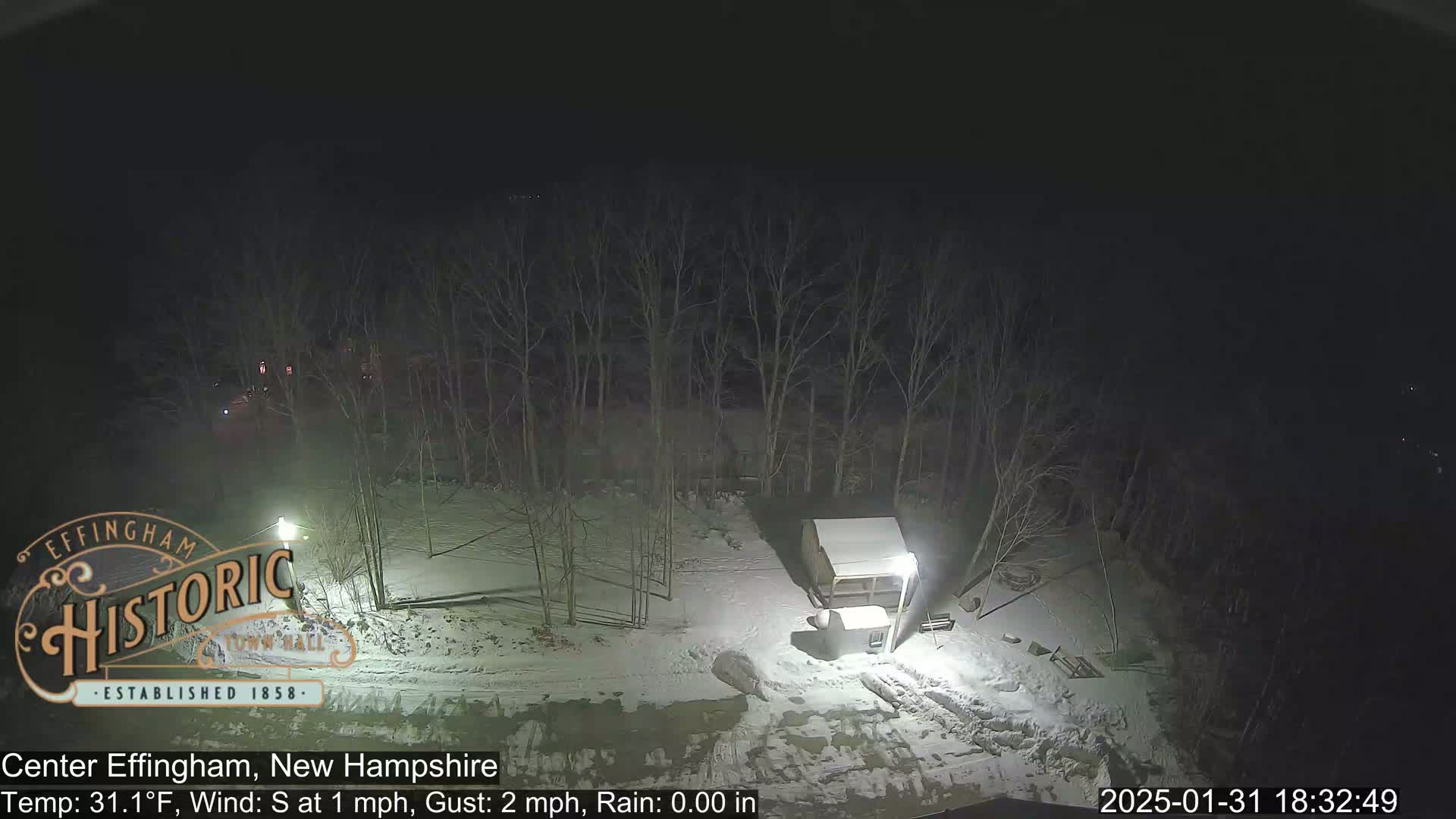 A nighttime, snowy scene shows a small structure and bare trees under a dark sky with light rain.