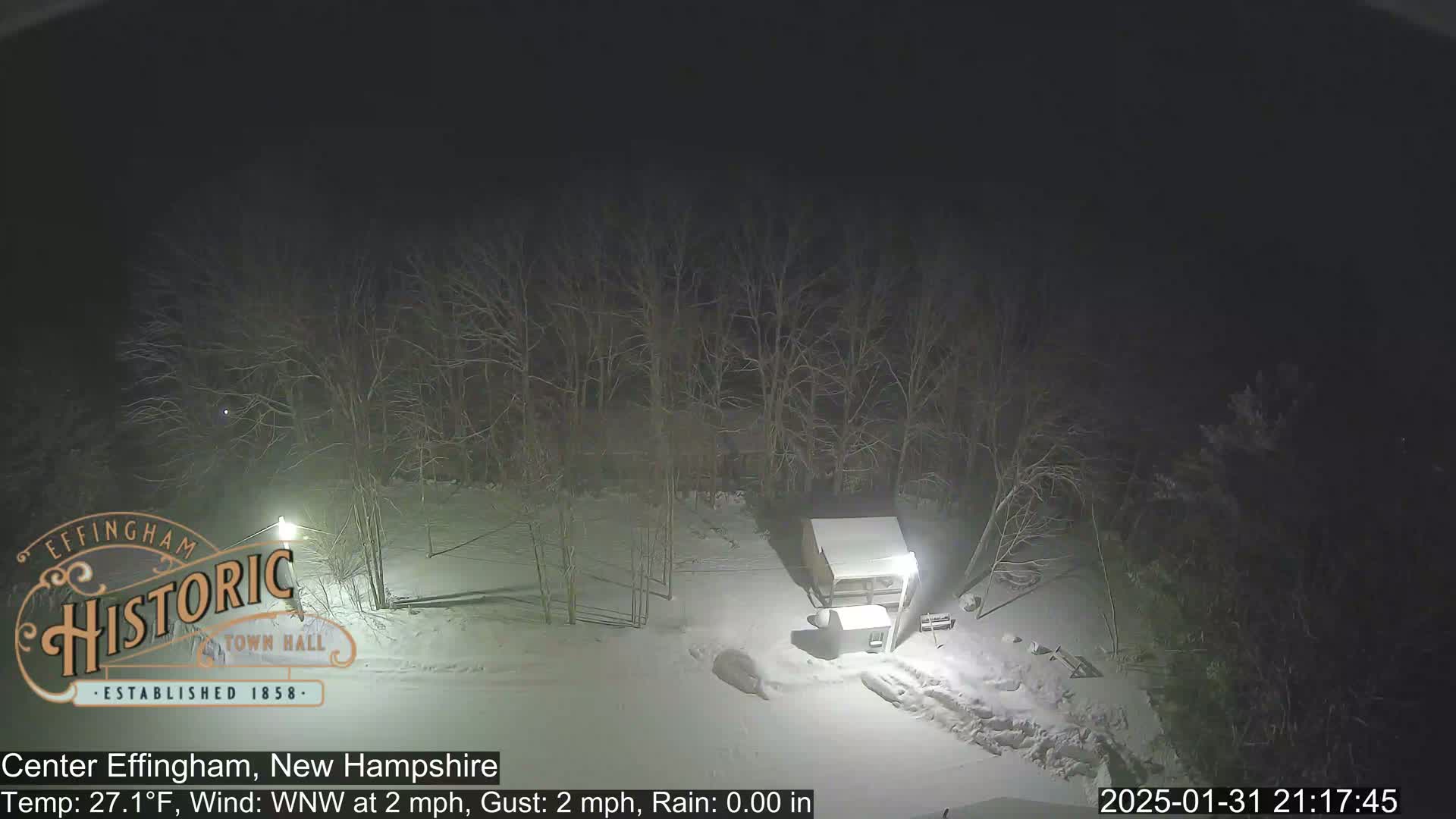A nighttime snow-covered scene shows a small building and bare trees under a dark sky.