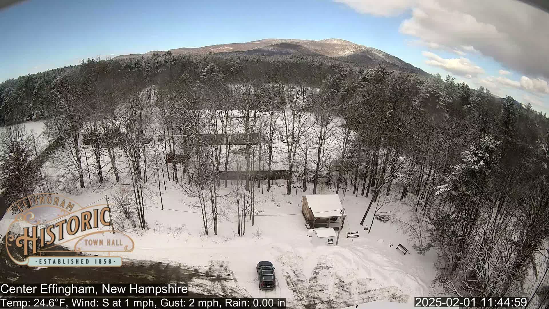 A snow-covered landscape with leafless trees, a small building, and a car in a parking area, all under a partly cloudy sky with mountains in the background.