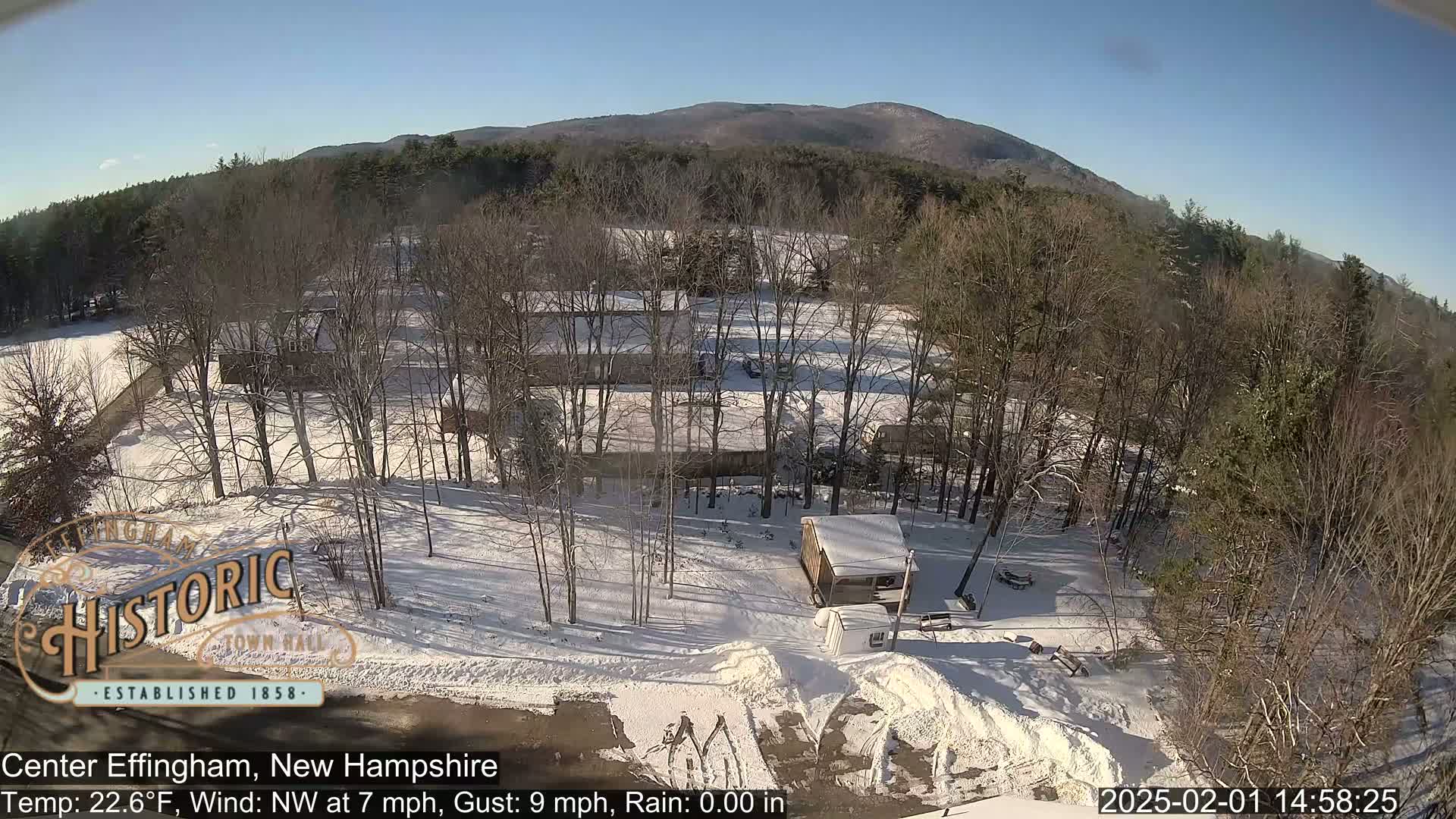 A snow-covered landscape with leafless trees and a small building is seen under a clear, sunny sky, with mountains visible in the background.
