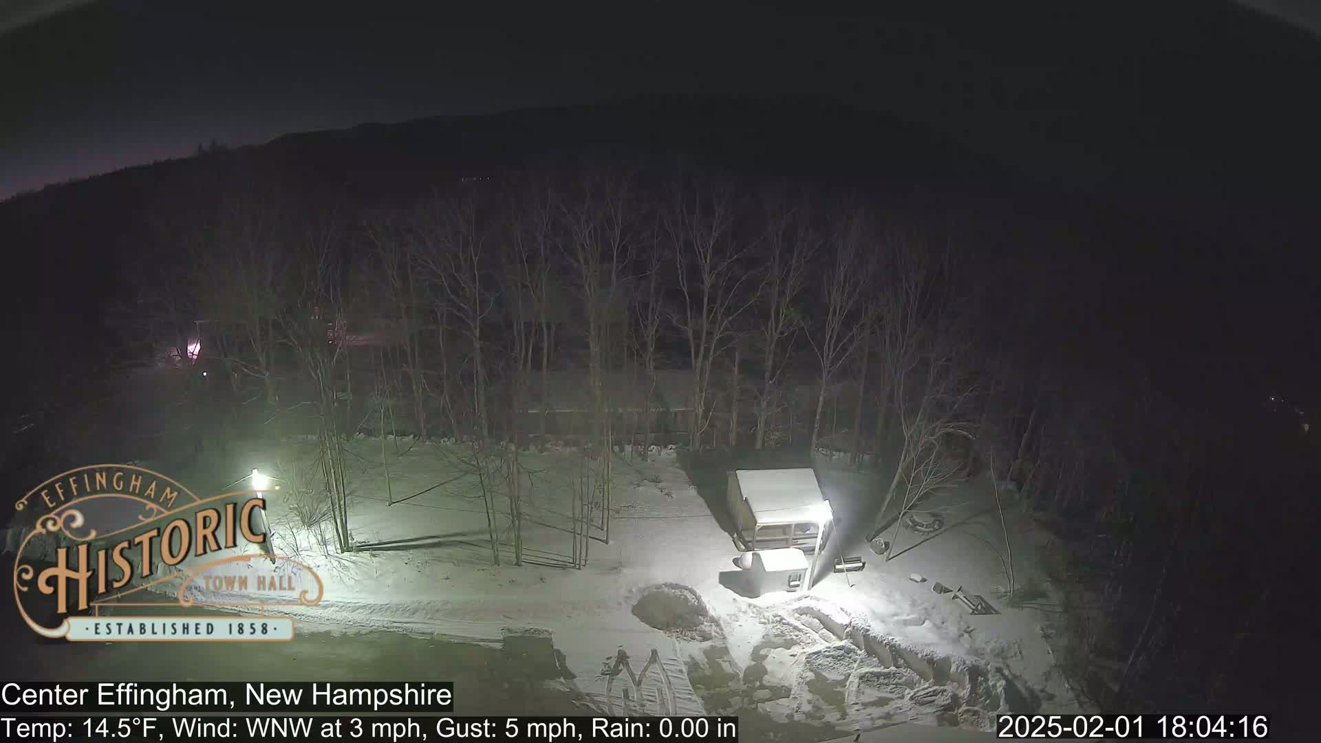 A nighttime, snow-covered scene shows leafless trees, a small building, and a partially lit area with what appears to be construction or excavation work.