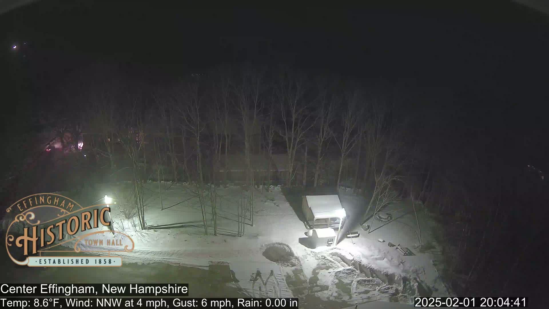 A nighttime, snow-covered scene shows a small building and bare trees under a dark sky.