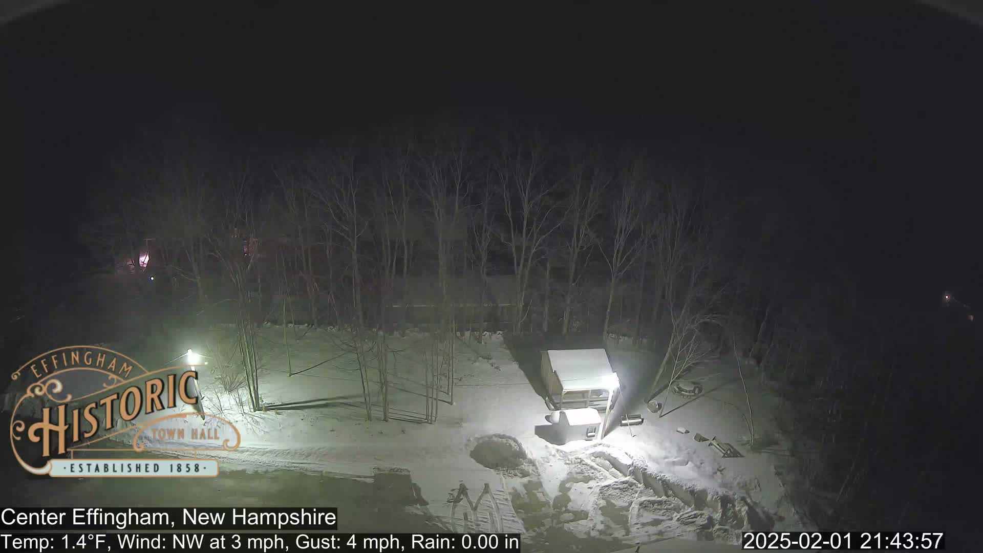 A nighttime, snow-covered scene shows a small shed and bare trees under a dark sky.
