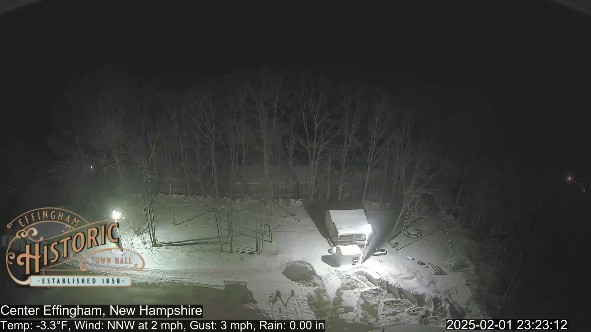 A nighttime, snow-covered scene shows a small shed and bare trees under a dark sky.