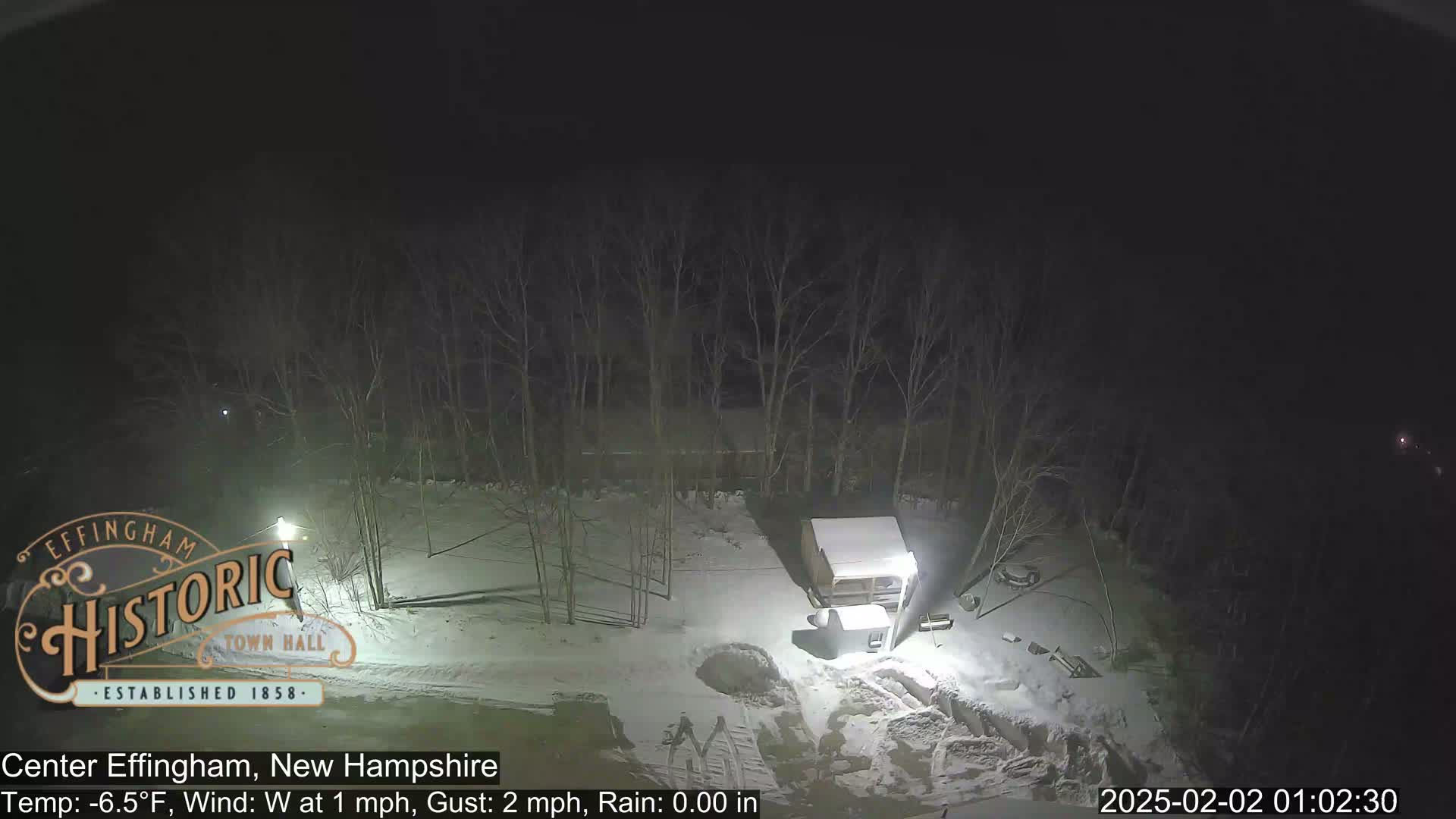 A nighttime, snow-covered scene shows a small building and bare trees under a dark sky with a slight westerly wind and a temperature of -6.5°F.