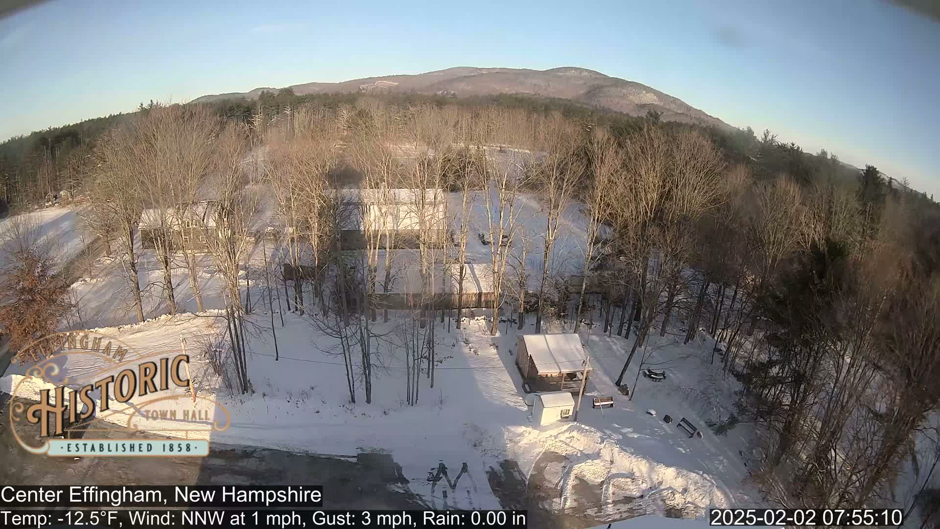 A snow-covered landscape with buildings and bare trees under a clear, sunny sky.
