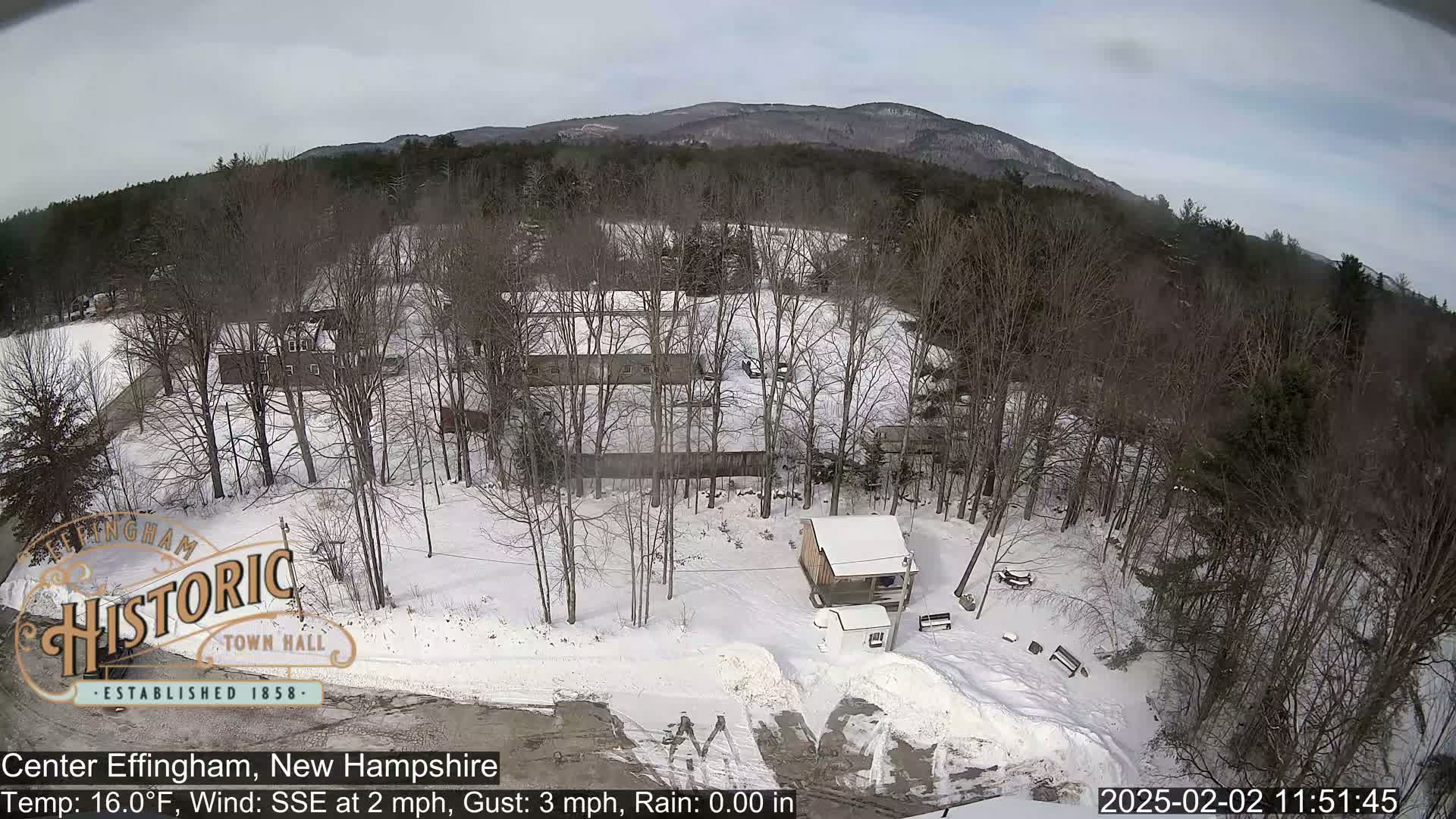 A snow-covered landscape with leafless trees, a small house, and distant mountains under a cloudy sky.