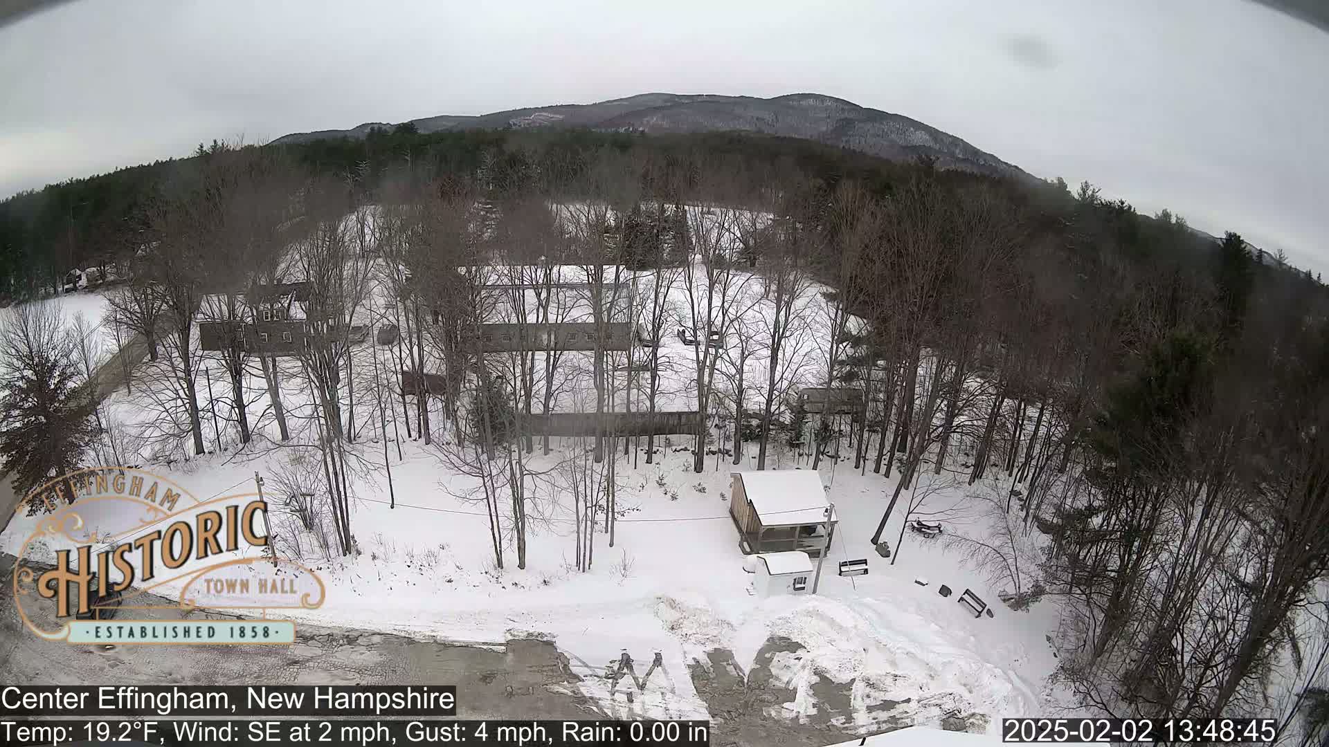 A snow-covered landscape with leafless trees, a small building, and distant mountains under a cloudy sky.