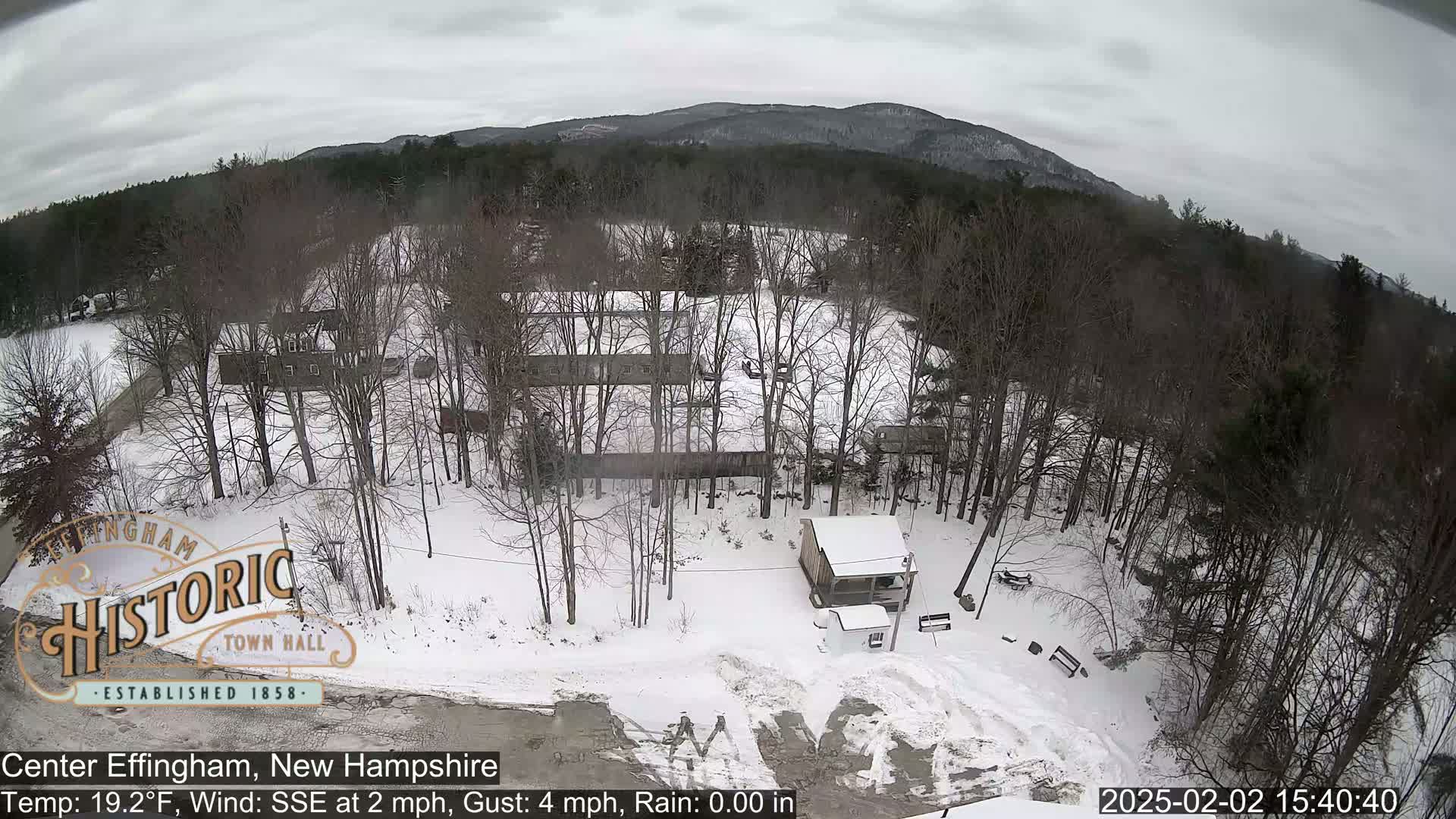 A snow-covered landscape with leafless trees, a small building, and distant mountains under an overcast sky.