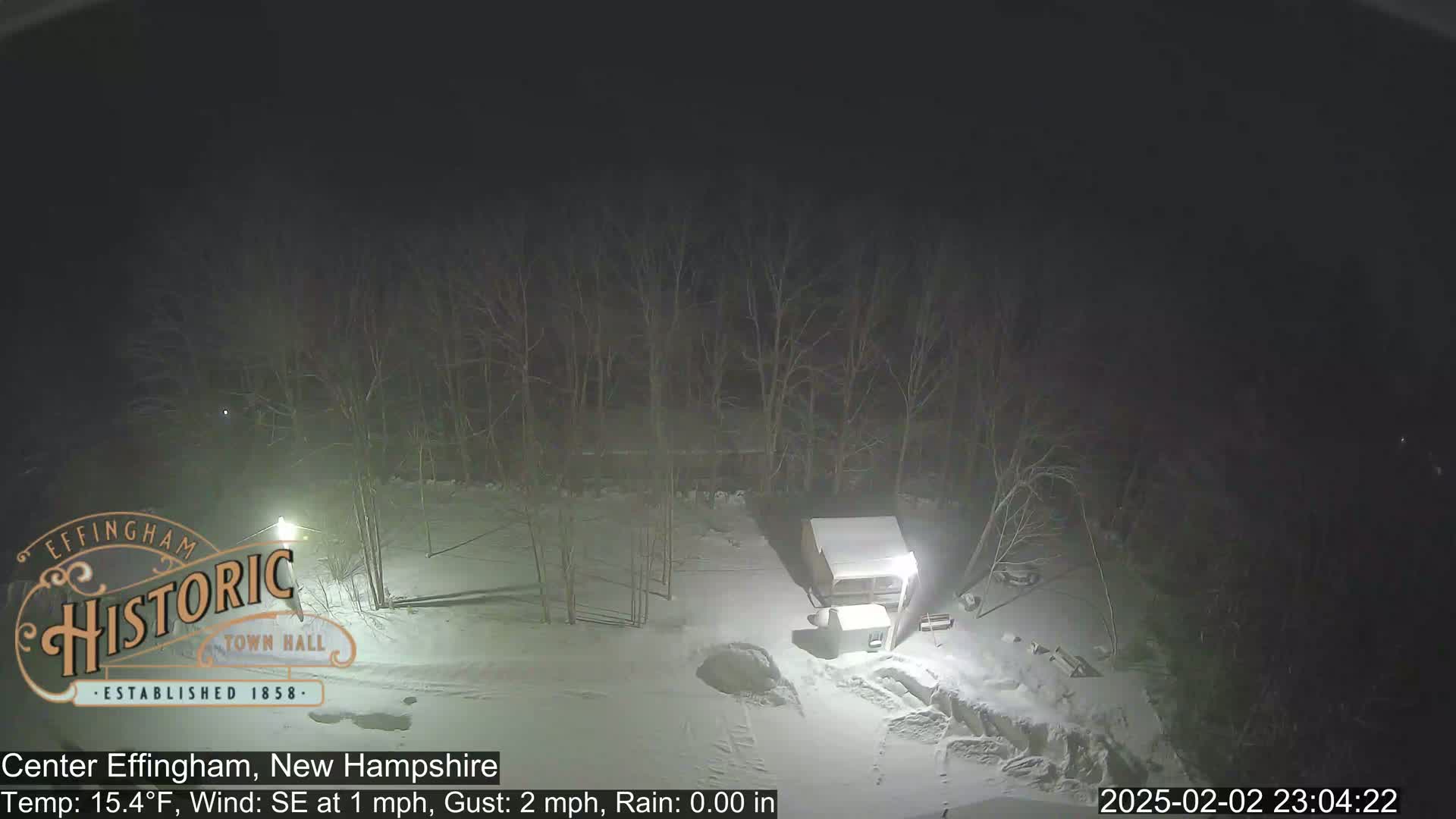 A nighttime, snowy scene shows a small structure and bare trees under a dark sky.