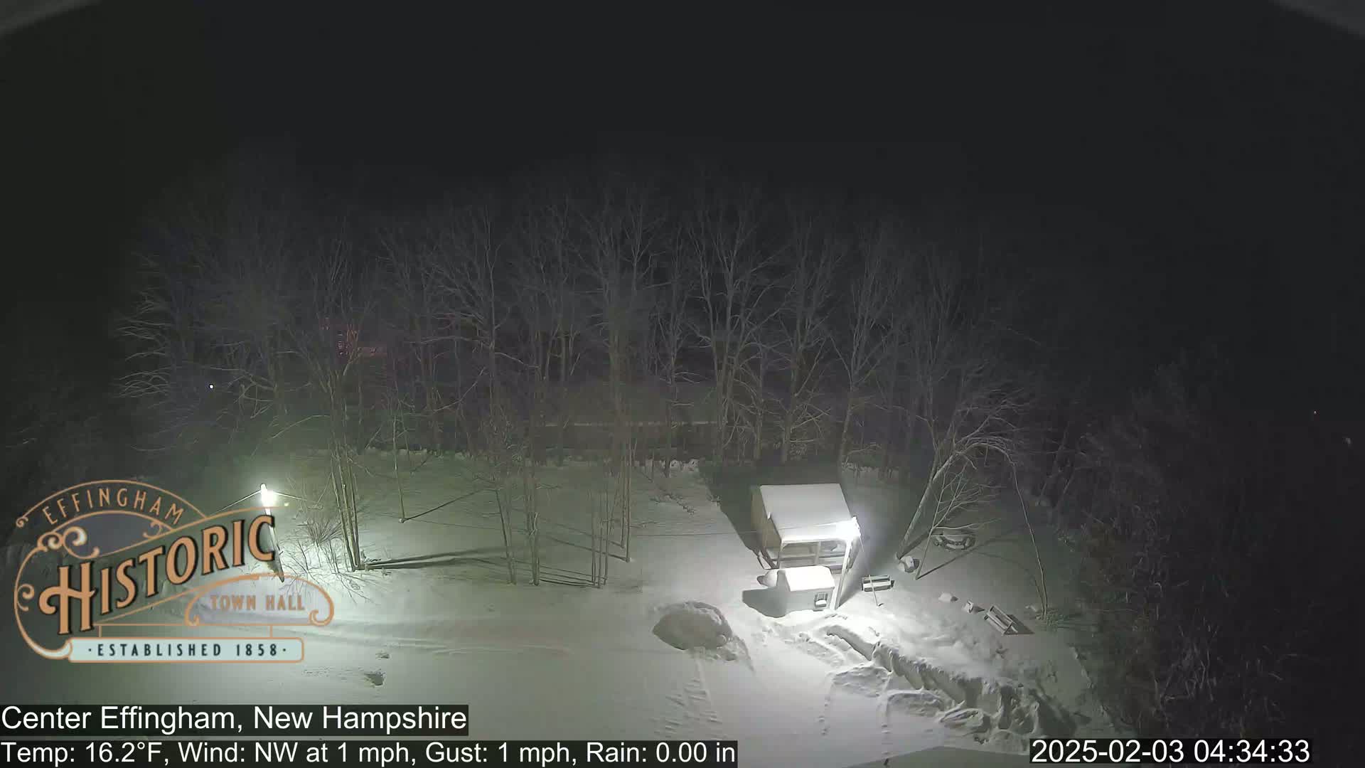 A snow-covered area at night, illuminated by lights, shows bare trees, a small structure, and a mostly clear sky with a light northwest wind.