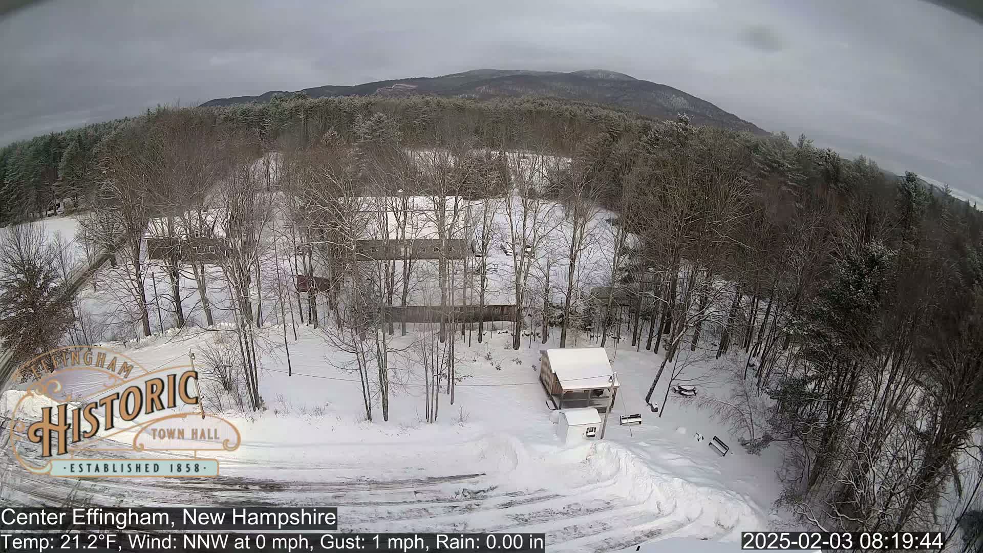 A snow-covered landscape with bare trees, a small building, and a road, viewed from above under an overcast sky.