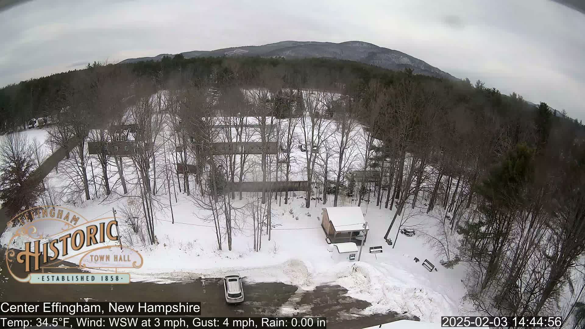 A snow-covered parking lot and surrounding wooded area with a small building are viewed from an elevated position, with mountains in the background under overcast skies.