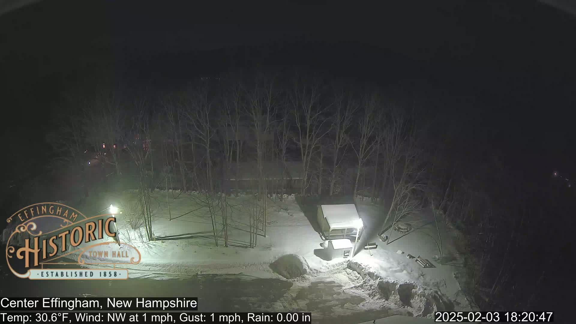 A nighttime, snow-covered scene shows a small structure and bare trees under a dark sky.
