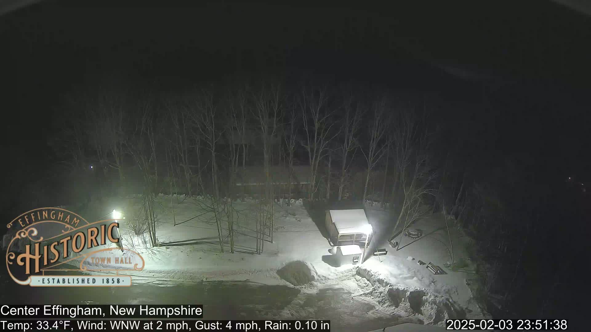 A nighttime, snowy scene shows a small building and a pile of snow under dim lighting, with bare trees in the background.