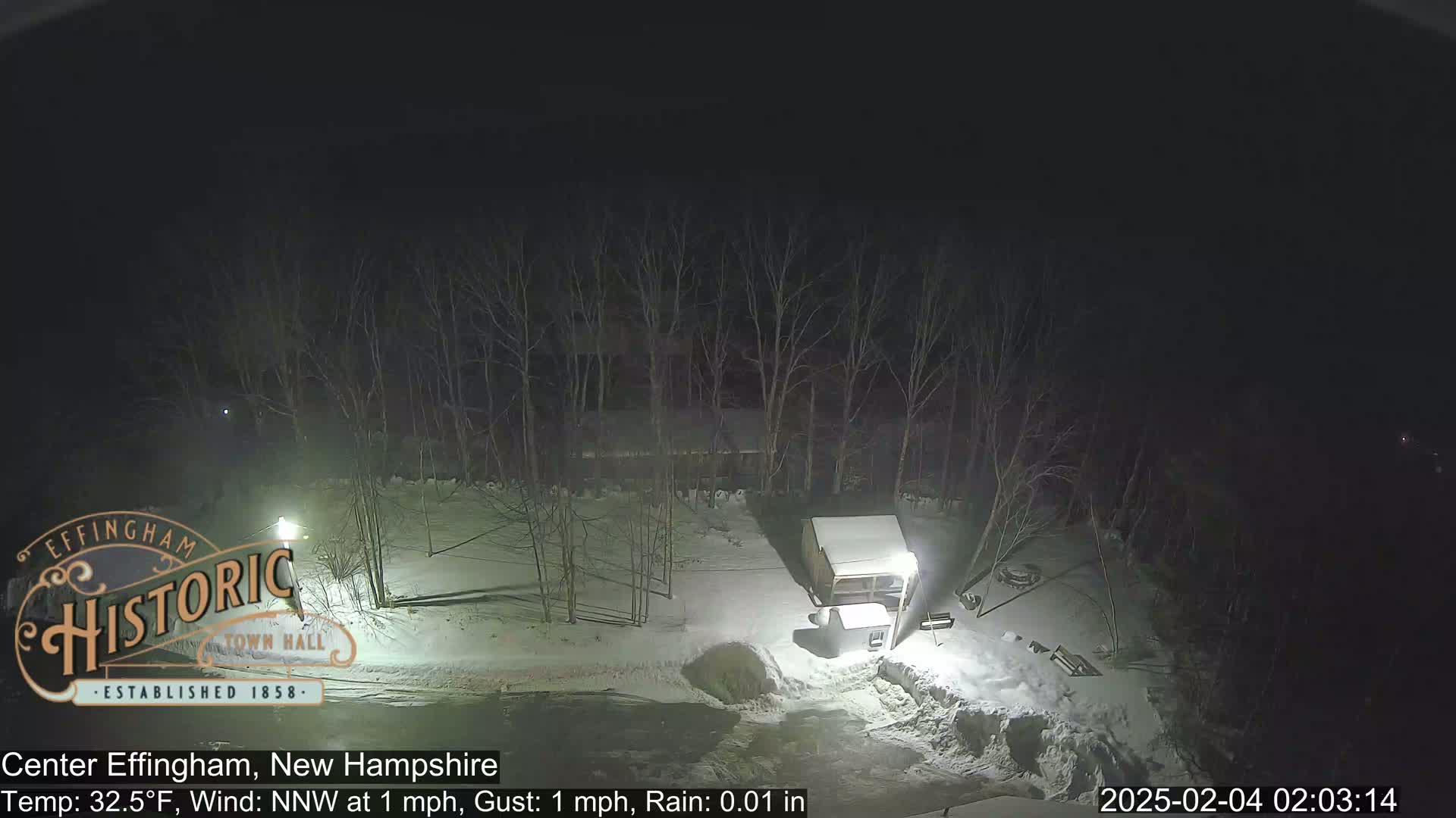 A nighttime, snowy scene shows a snow-covered yard with a small structure and bare trees under a dark sky, with light rain.