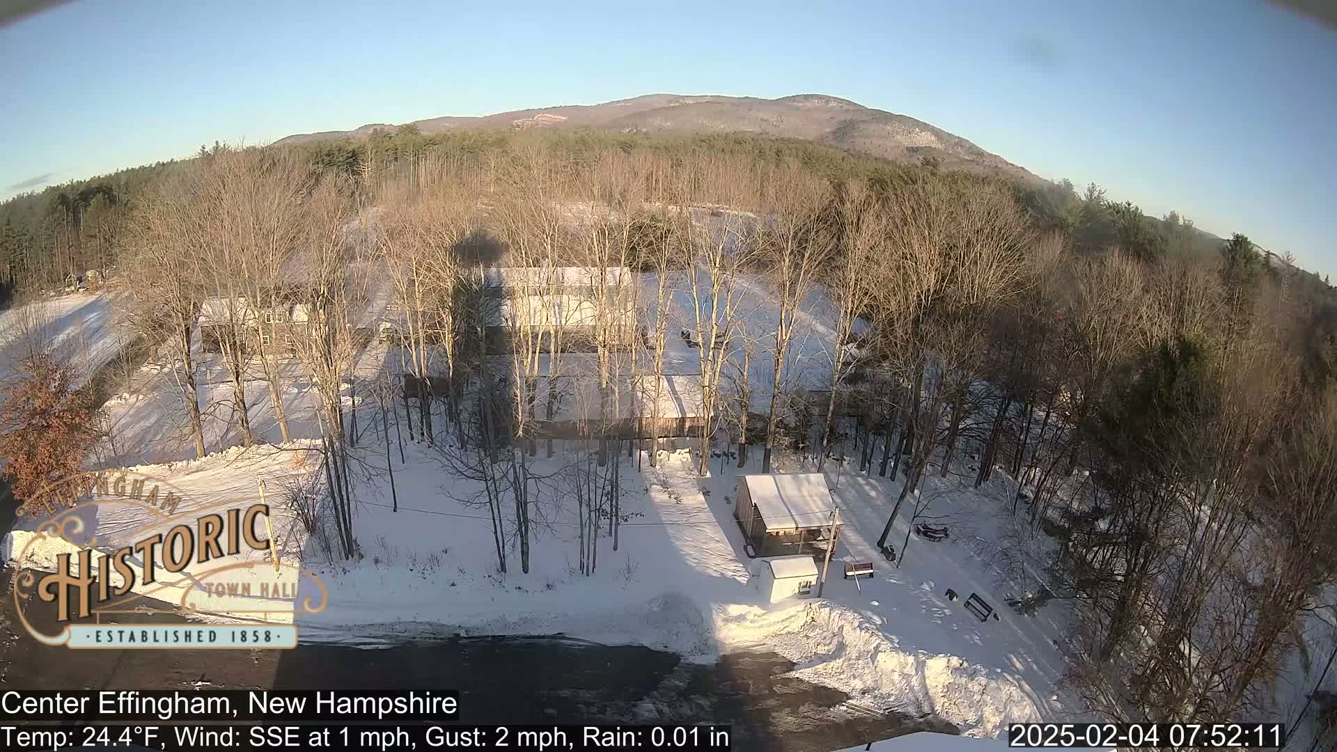 A snow-covered landscape with bare trees, a few buildings, and a mountain in the background under a clear, sunny sky.