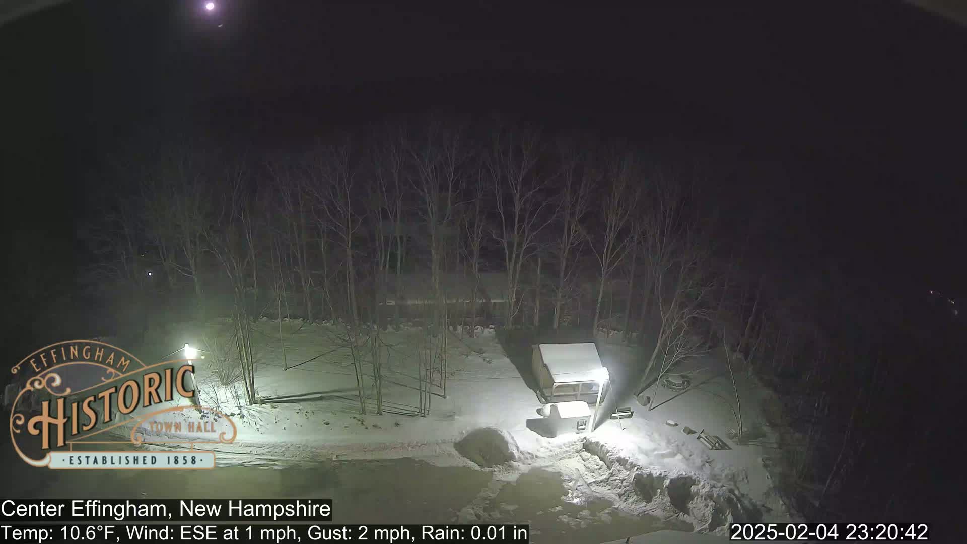 A nighttime, snowy scene shows a small building and bare trees under a partly cloudy sky with light rain.