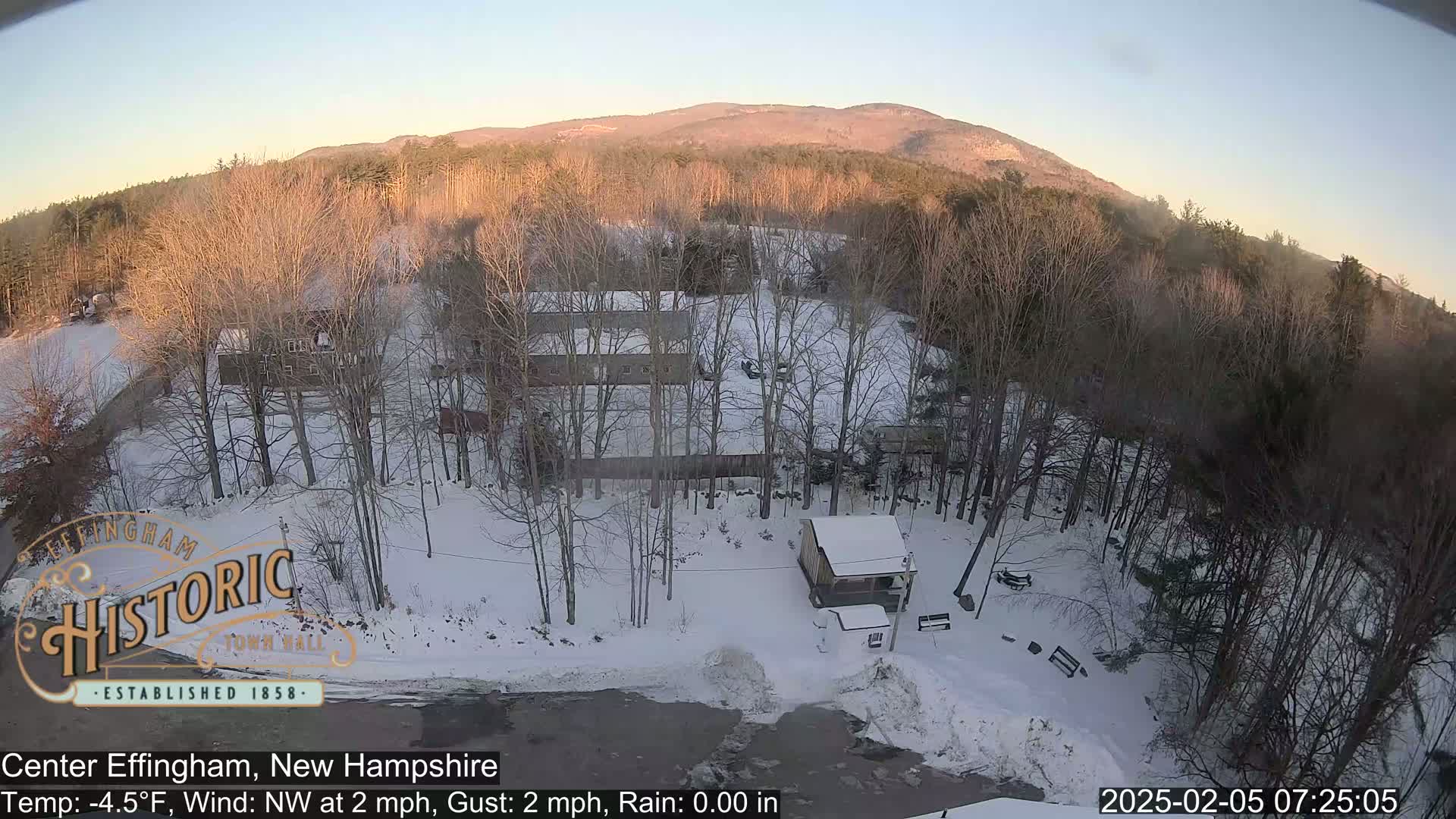 A snow-covered landscape with bare trees and a few buildings, viewed from above in the early morning light, under a clear sky.