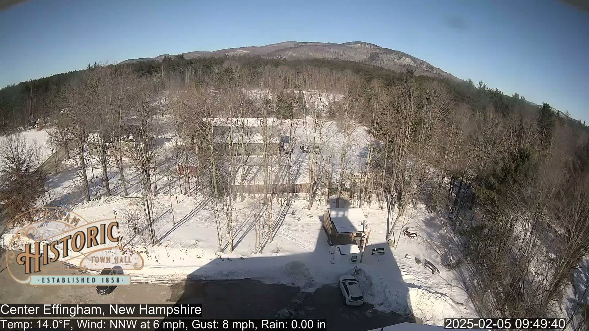 A snow-covered landscape with leafless trees, a few buildings, and a car in a parking lot under a clear, sunny sky.