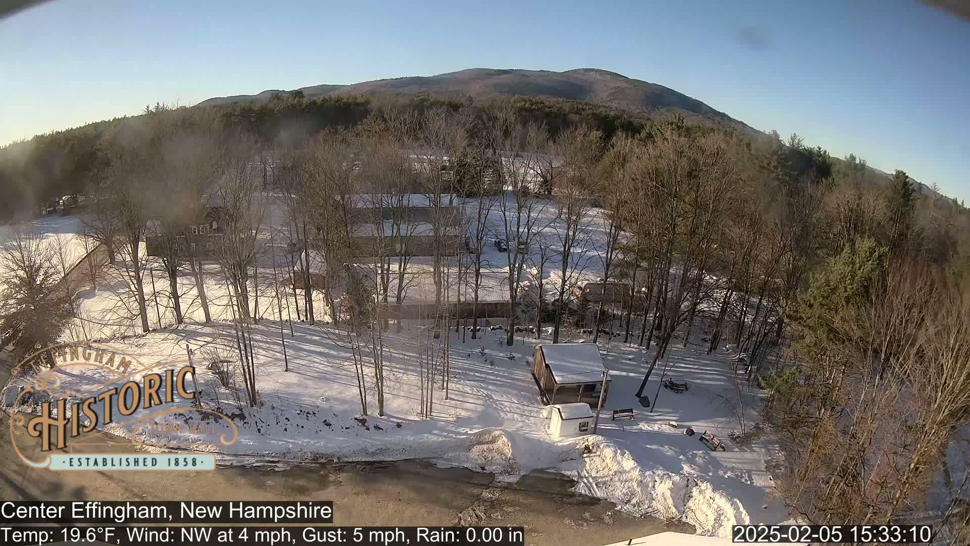 A snow-covered landscape with leafless trees, a small cabin, and buildings, viewed from above on a sunny day.