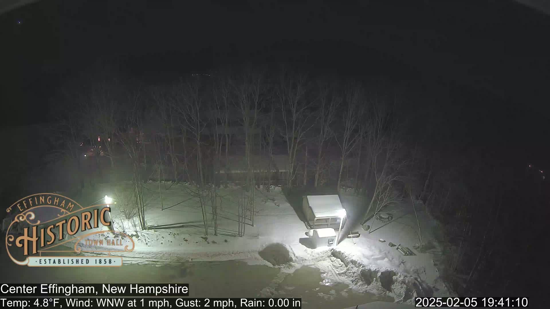 A nighttime, snowy scene shows a small shed and bare trees under a dark sky with light rain.