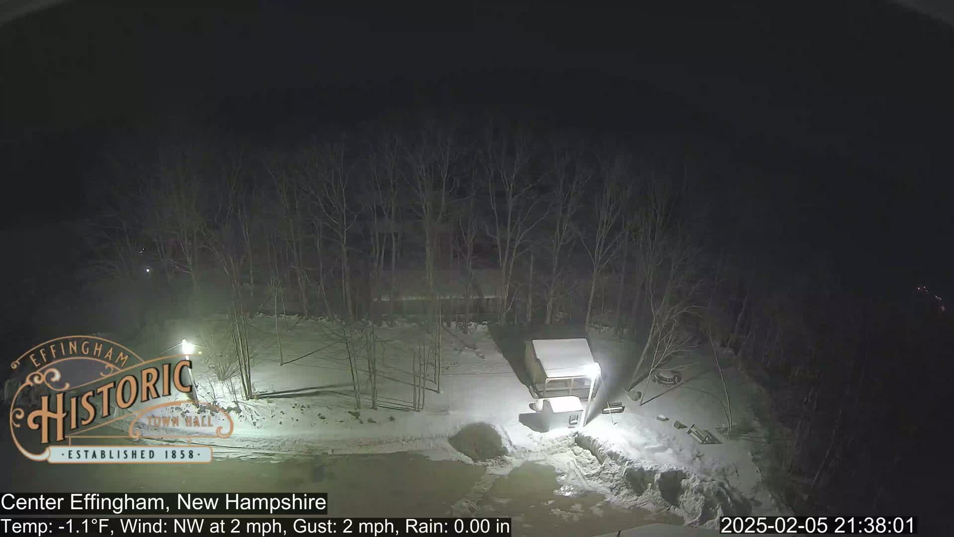 A snow-covered, nighttime scene shows bare trees, a small shed, and some outdoor lights.