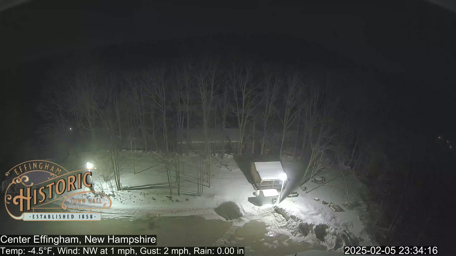 A nighttime, snowy scene shows a small building and bare trees under a dark sky.