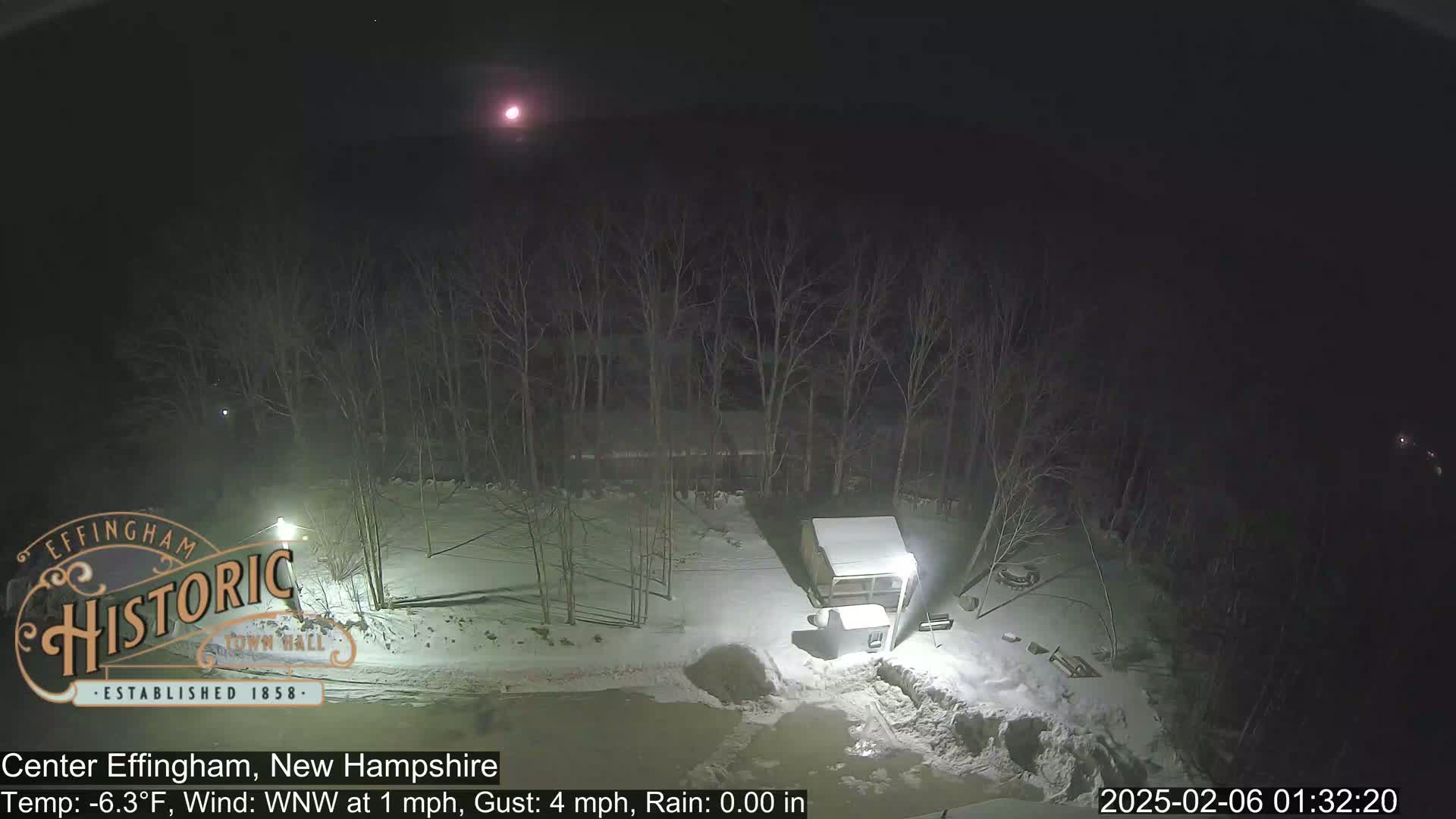 A nighttime, snowy scene shows a small, snow-covered structure and bare trees under a partly cloudy sky illuminated by a bright moon.