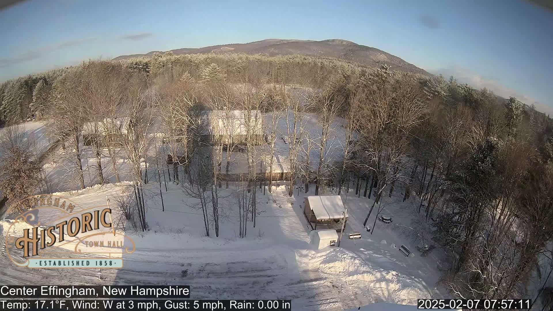 A snow-covered landscape with buildings and leafless trees under a clear, sunny sky.