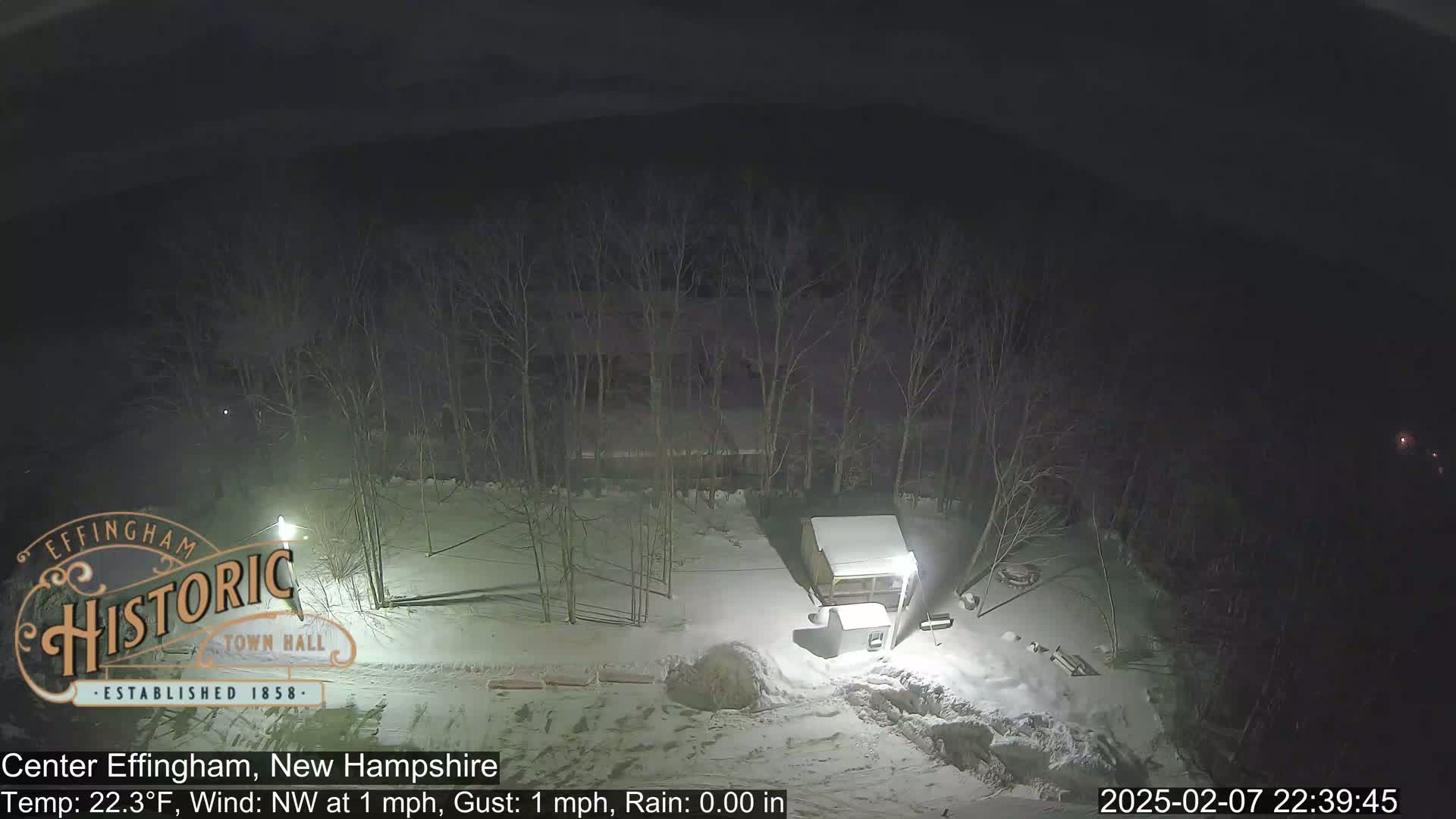 A nighttime snow-covered scene shows a small building and bare trees under a dark sky.