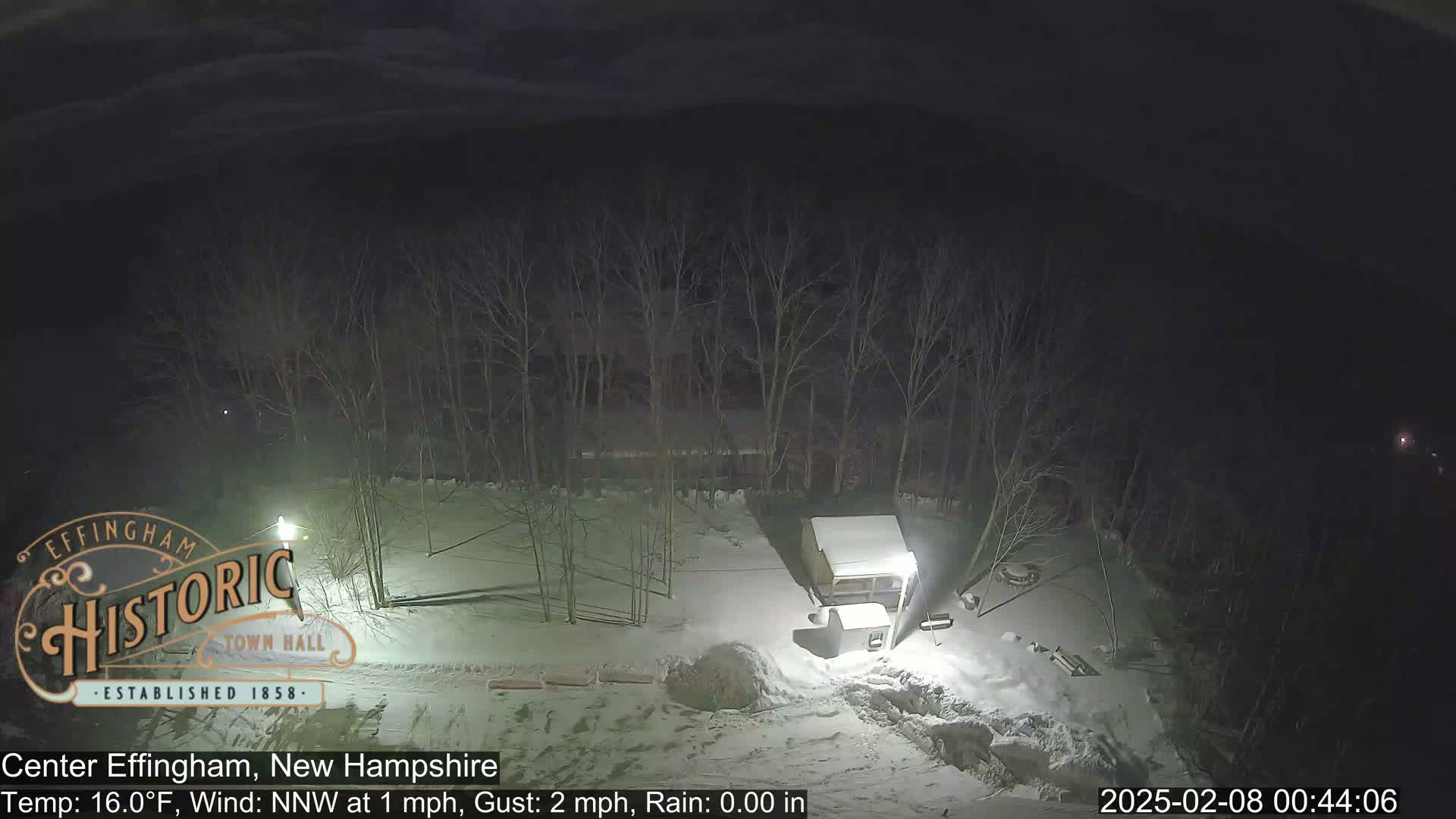 A snow-covered nighttime scene shows a small shed and bare trees under a dark sky with a light wind.