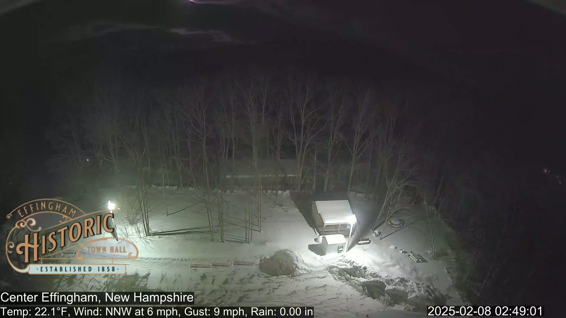 A nighttime, snow-covered scene shows a small building and bare trees under a dark sky.