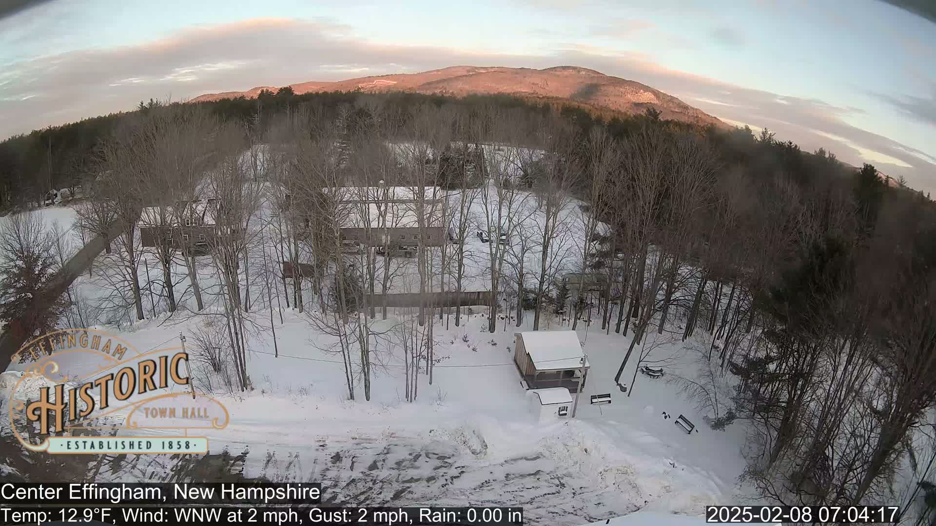 A snow-covered landscape with bare trees and several buildings, viewed from above under a partly clear sky at sunrise.