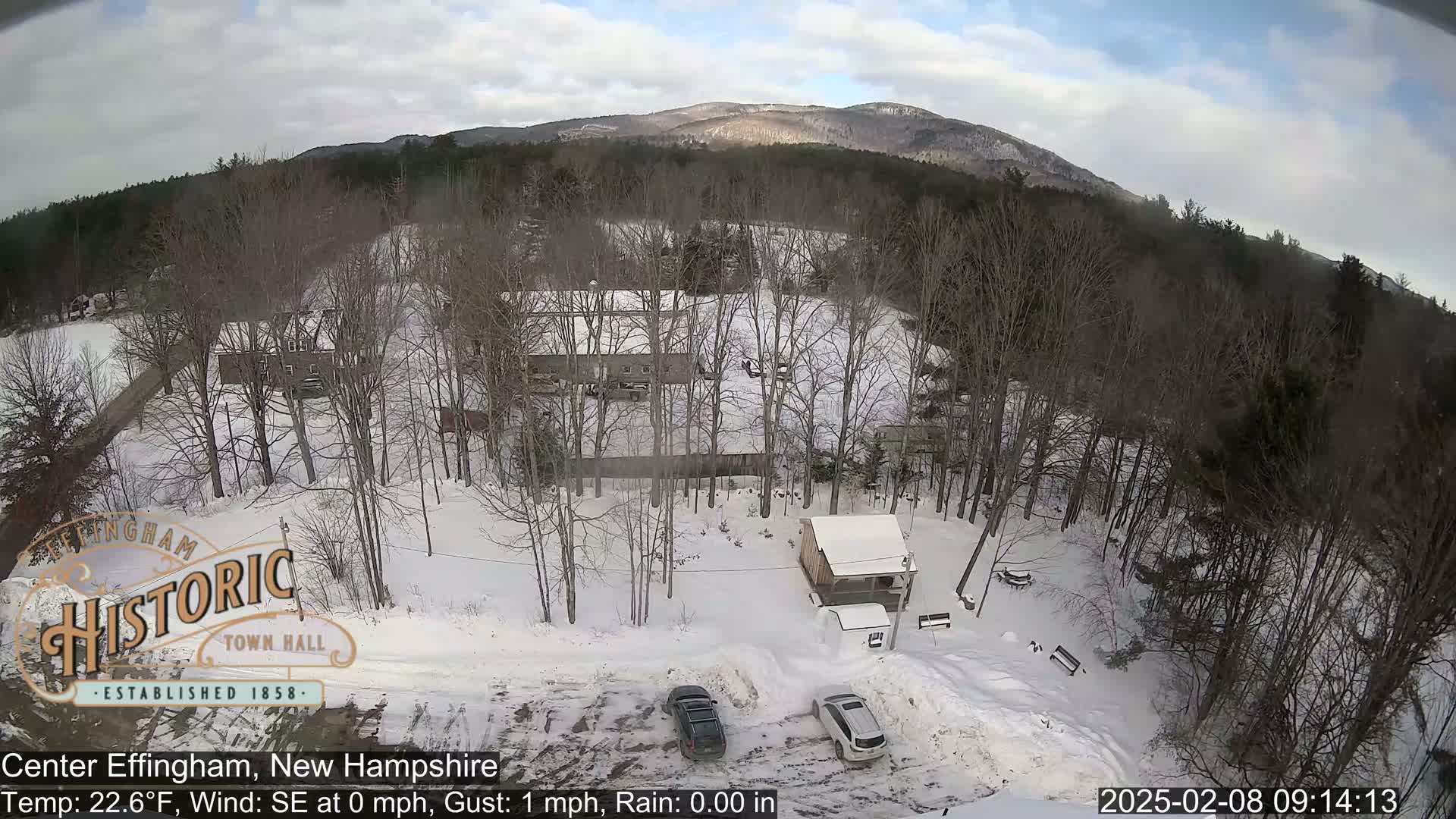 A snow-covered landscape with bare trees, buildings, and parked cars is visible under a partly cloudy sky.