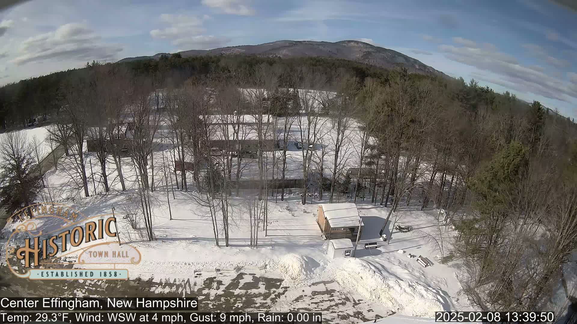A snow-covered landscape with leafless trees, a small building, and a mountain in the background under a partly sunny sky.