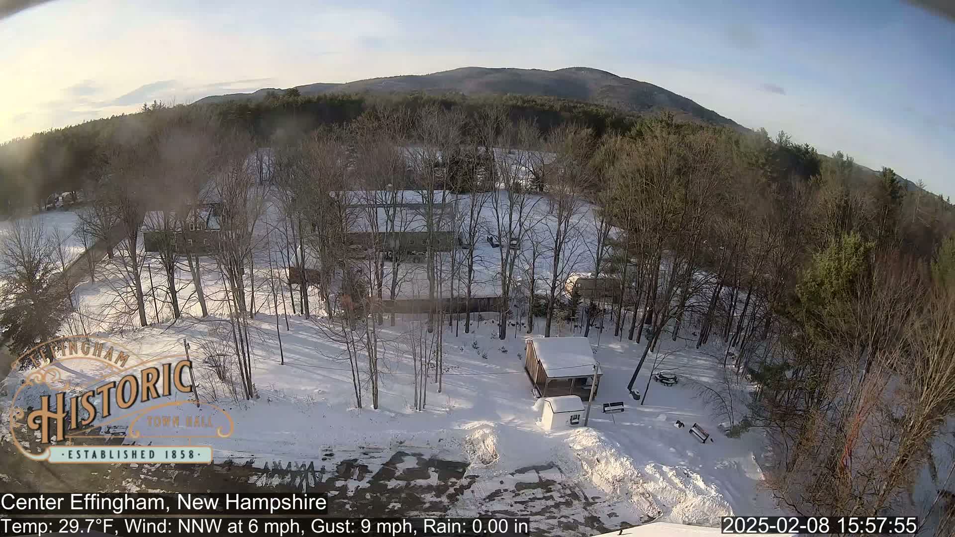 A snow-covered landscape with leafless trees, a small building, and a distant mountain range under a mostly clear sky.