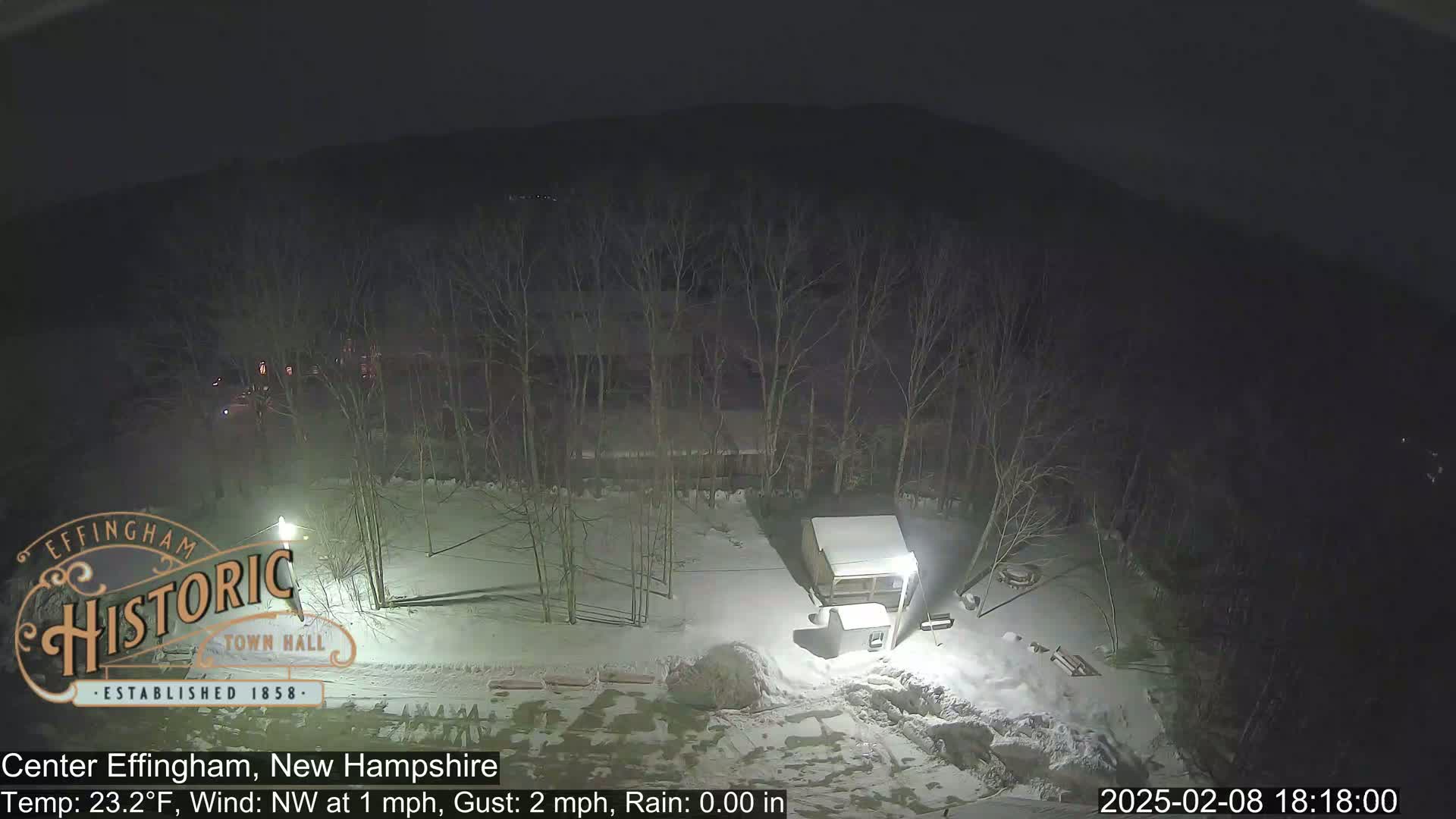 A nighttime, snow-covered scene shows a small building and bare trees under a dark sky.