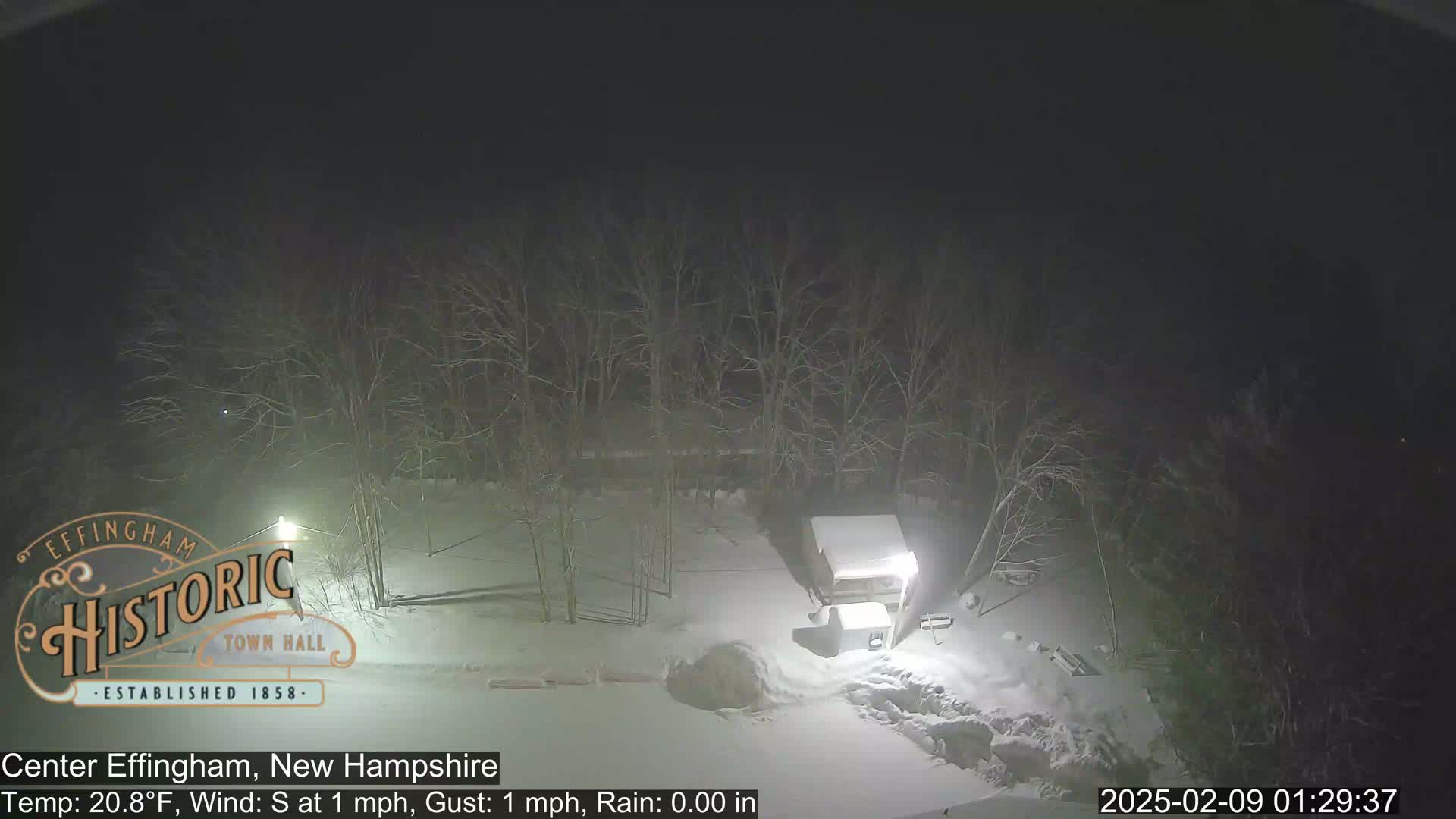 A nighttime, snowy scene shows a snow-covered shed and bare trees under a dark sky with light rain.