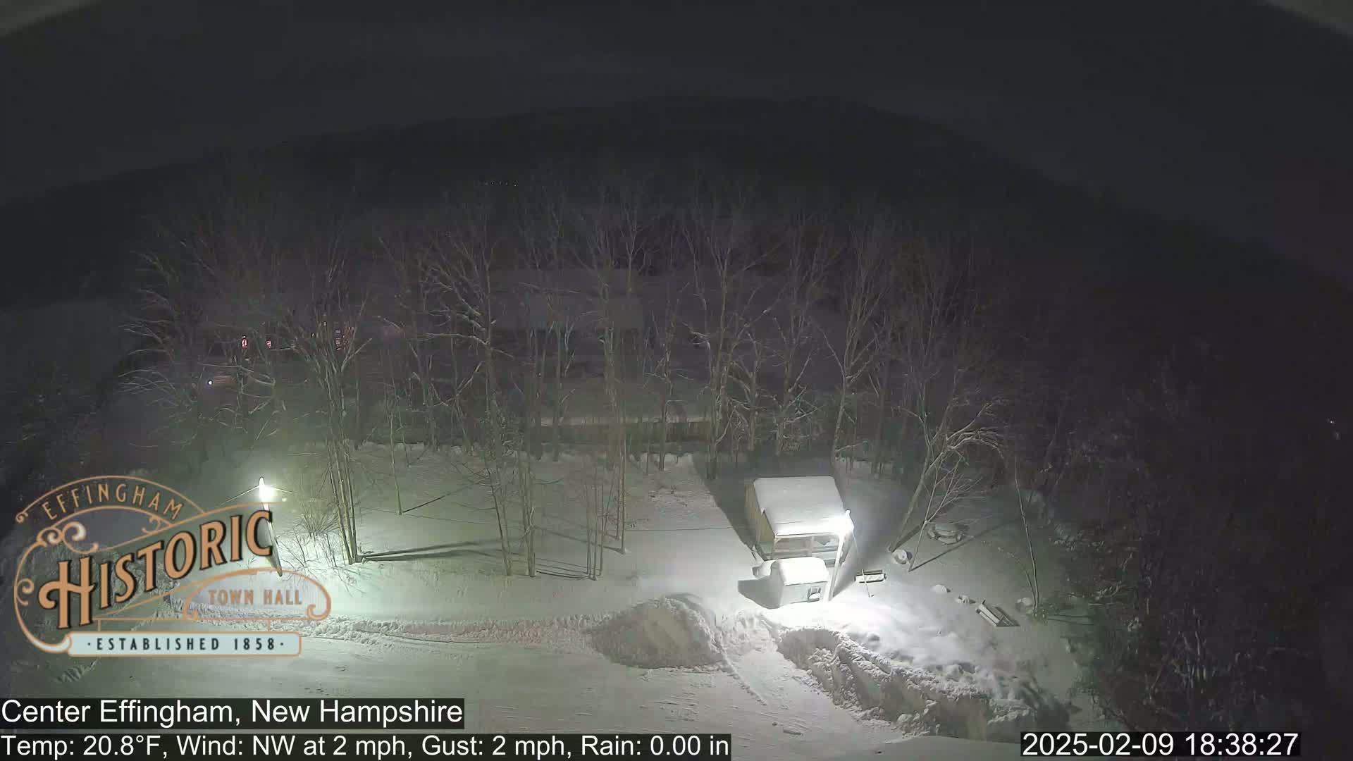 A nighttime, snow-covered scene shows leafless trees surrounding a small building, under a dark sky.