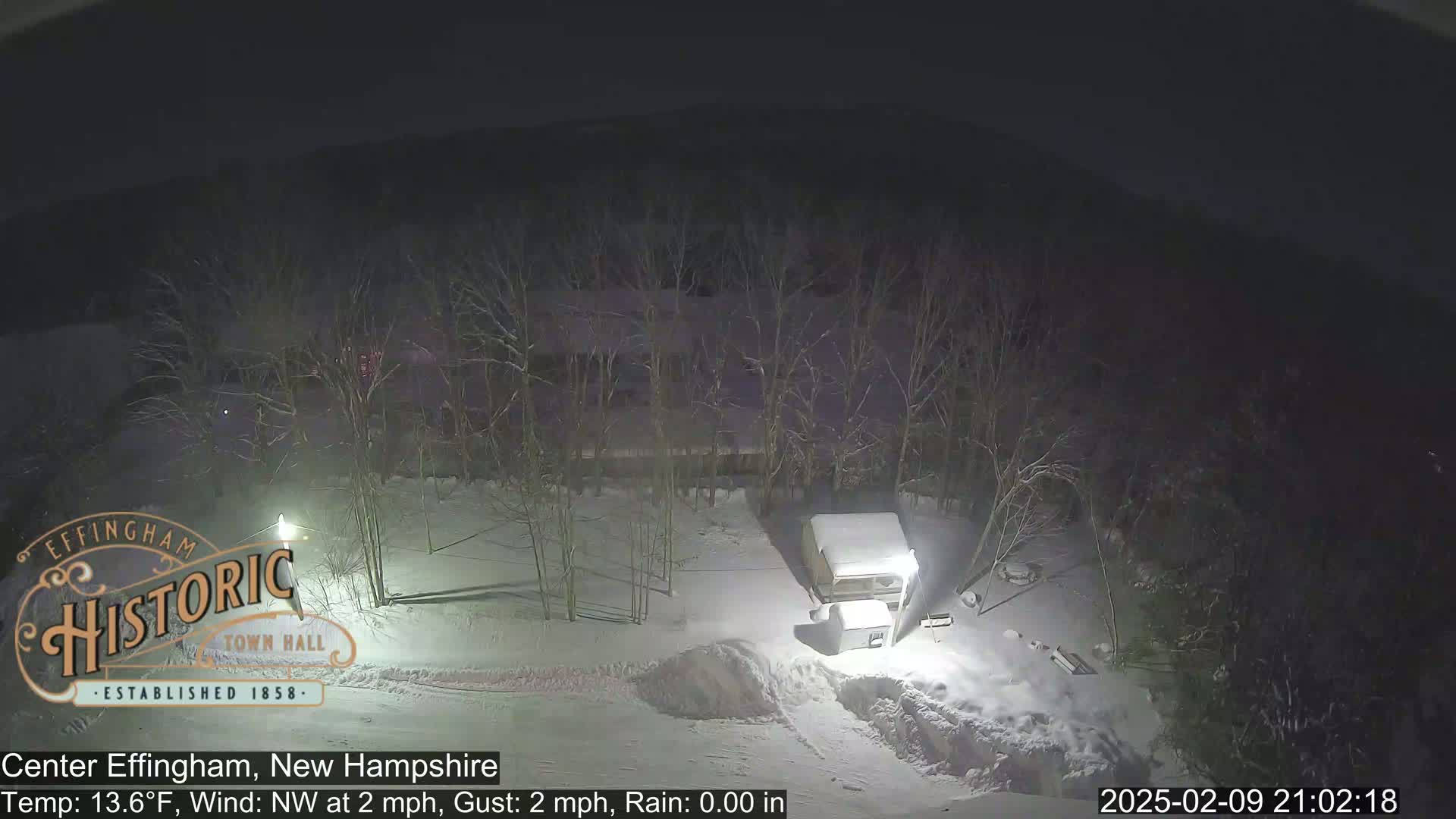 A snow-covered area at night, with leafless trees and a snow-covered vehicle visible, under a dark sky.